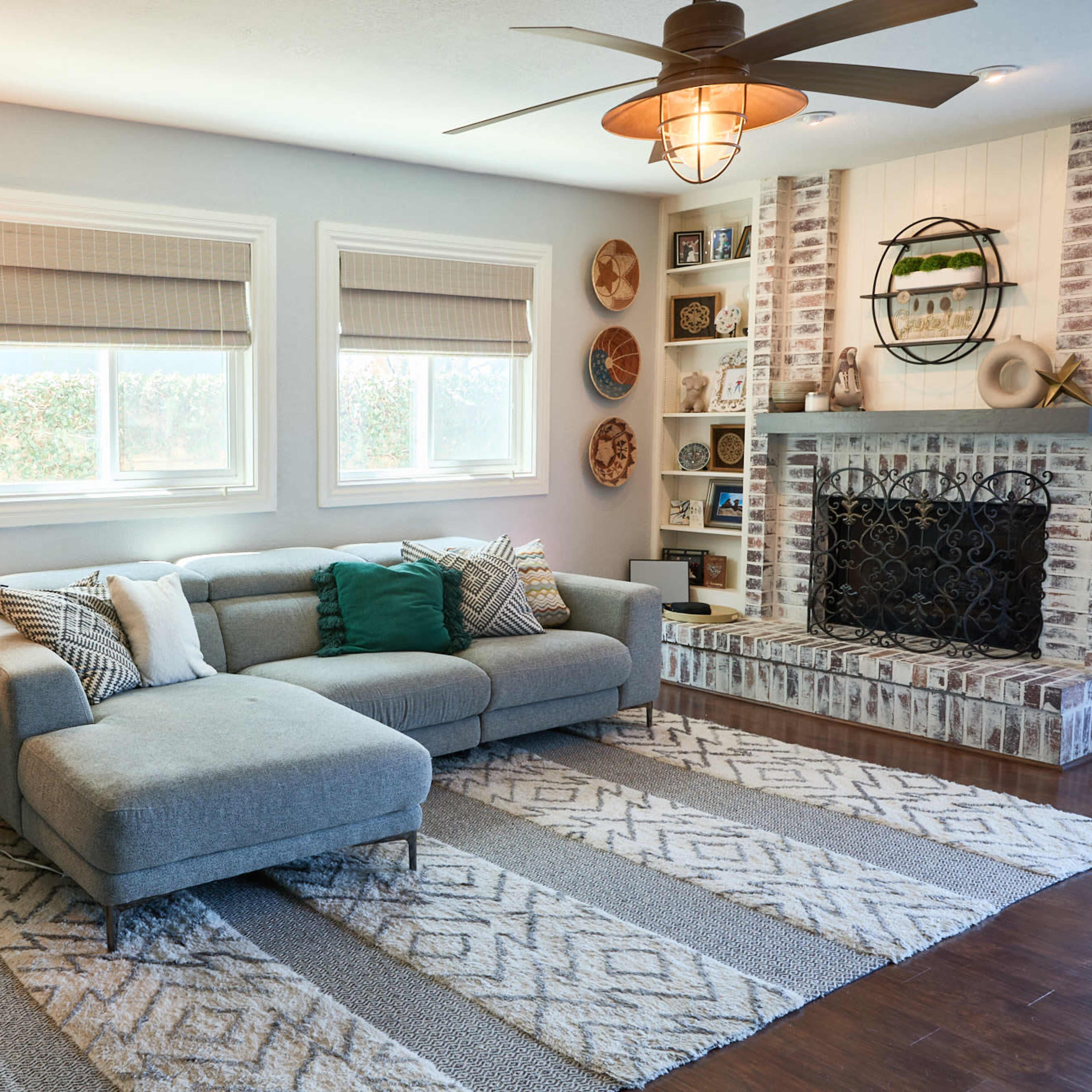 The living room features a gray sectional sofa, a decorative rug, and a brick fireplace with shelves, illuminated by a ceiling fan and natural light from two windows.