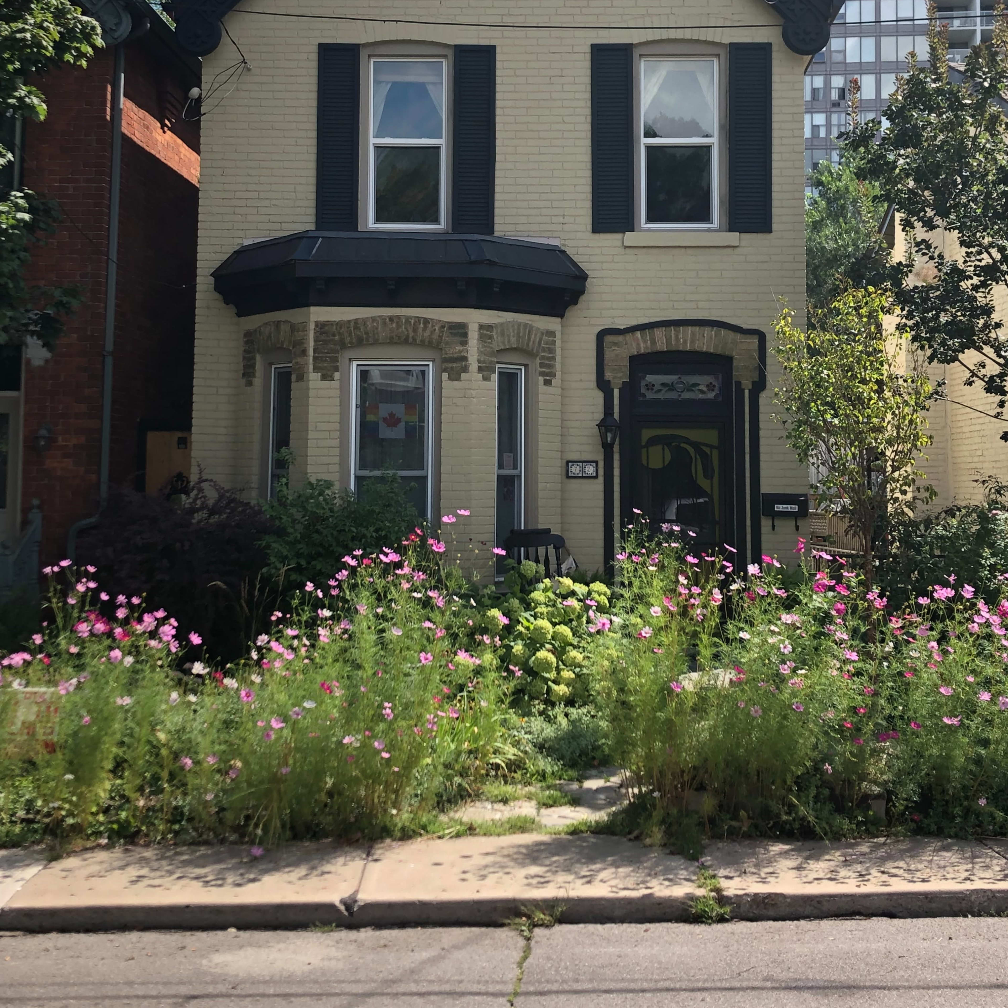 A two-story yellow brick house with black trim has a flower garden in front, featuring pink blooms, and is located next to a tall apartment building.