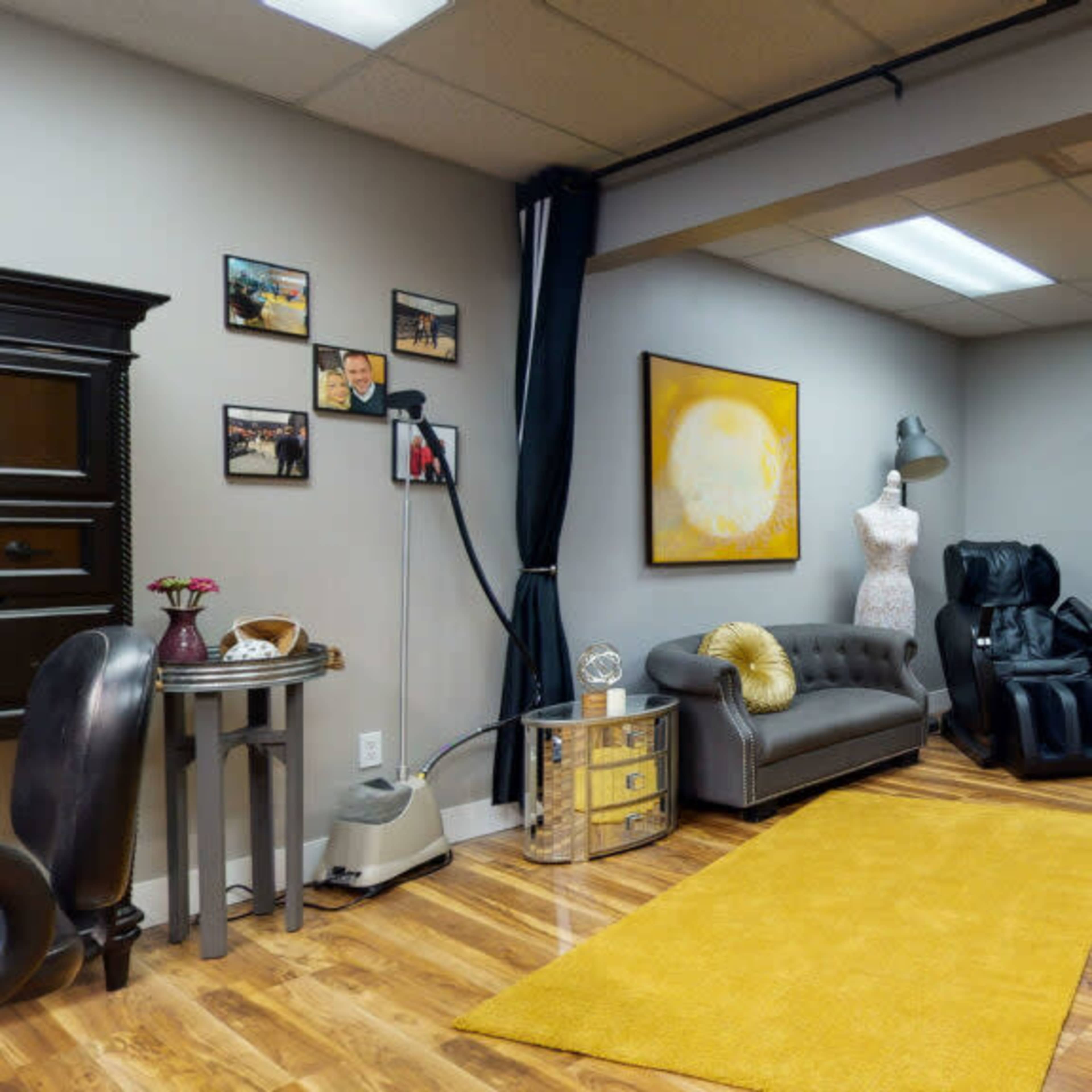 A spacious room with hardwood flooring, featuring a gray couch, a mannequin, and a clothing rack, all arranged around a central yellow area rug.