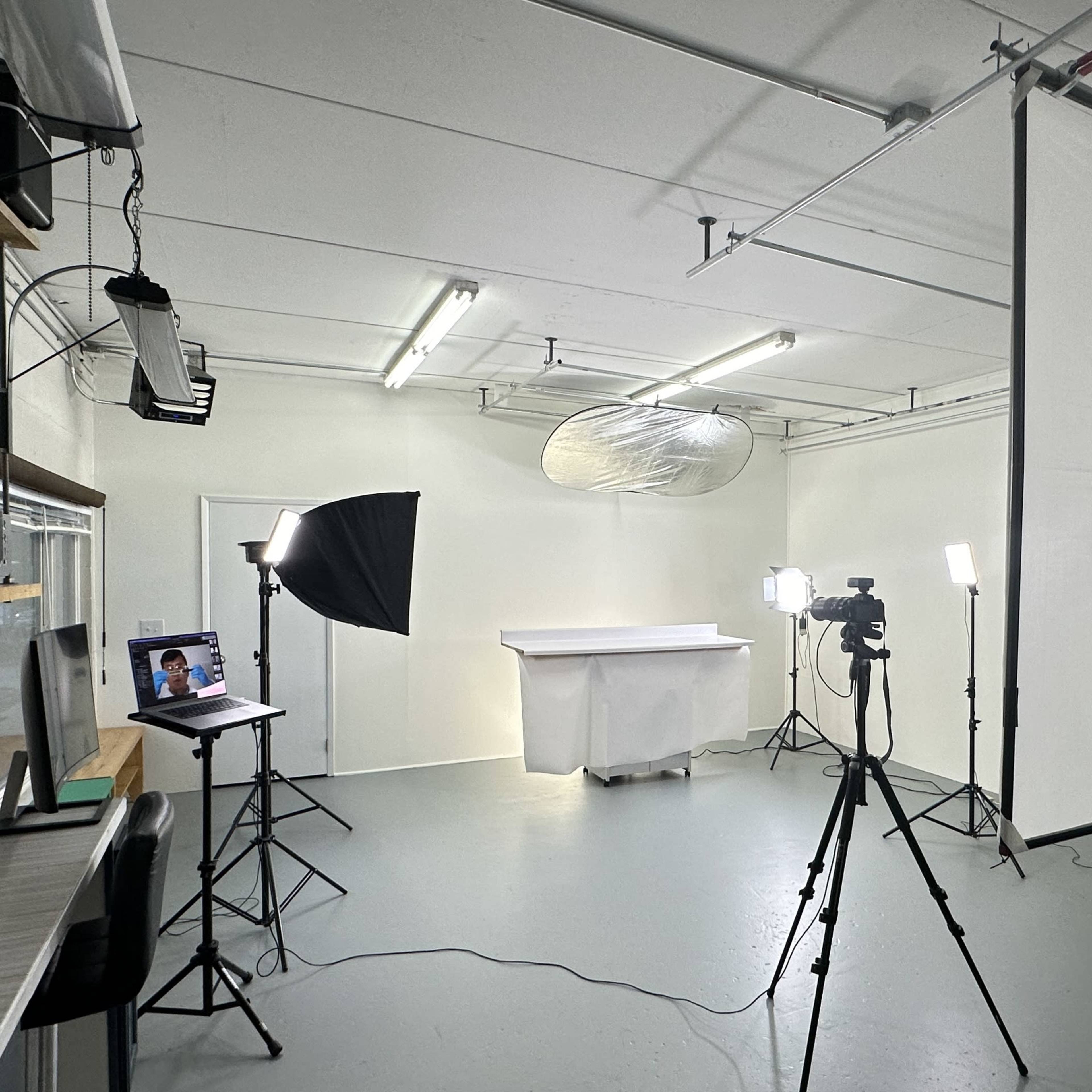 A photo studio setup features a white backdrop, multiple softbox lights, two cameras on tripods, and a table covered with a cloth.