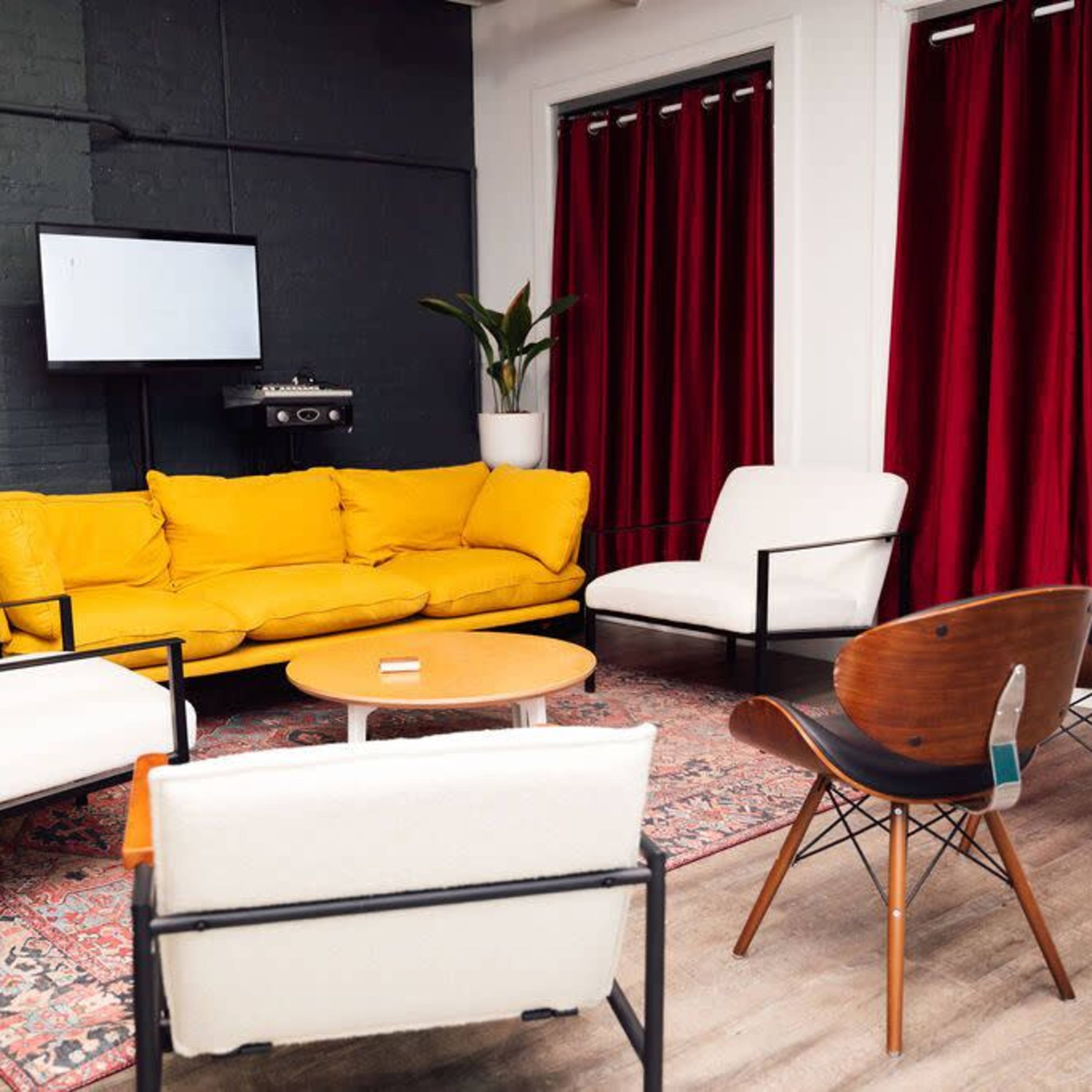 A modern seating area with a bright yellow couch, white chairs, a round table, and red curtains in a room featuring plants and a television on the wall.