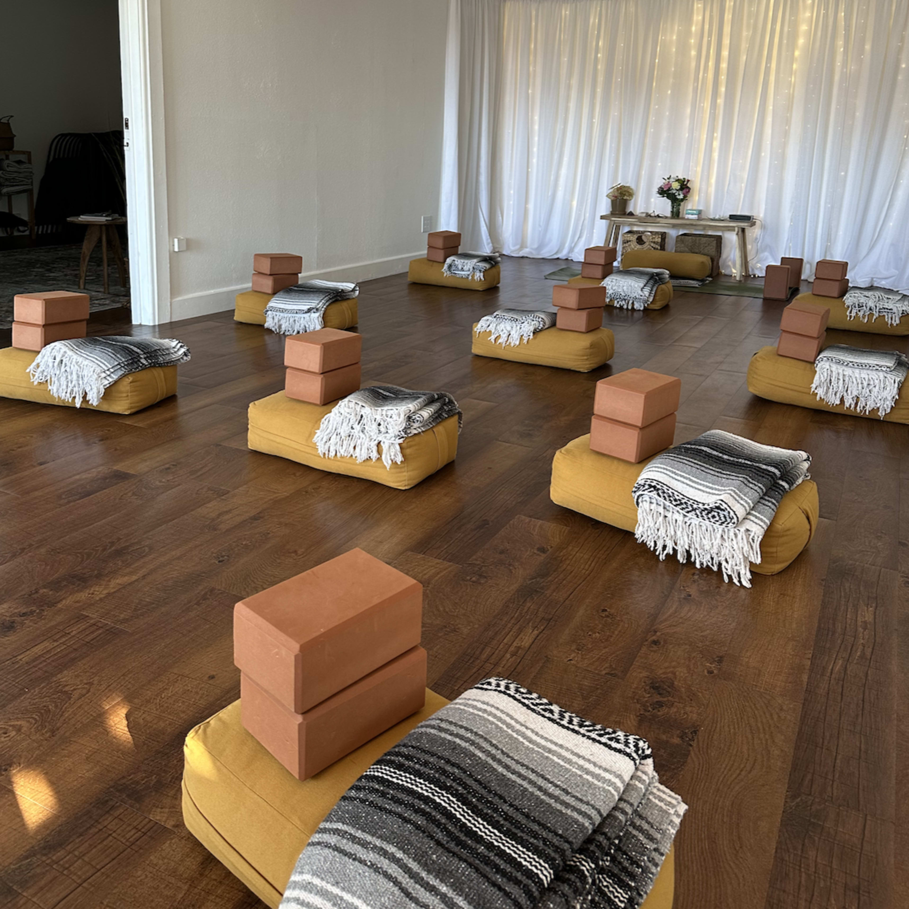A room arranged for a yoga or meditation session, featuring mustard-colored seating cushions, yoga blocks, and blankets neatly organized on a wooden floor.