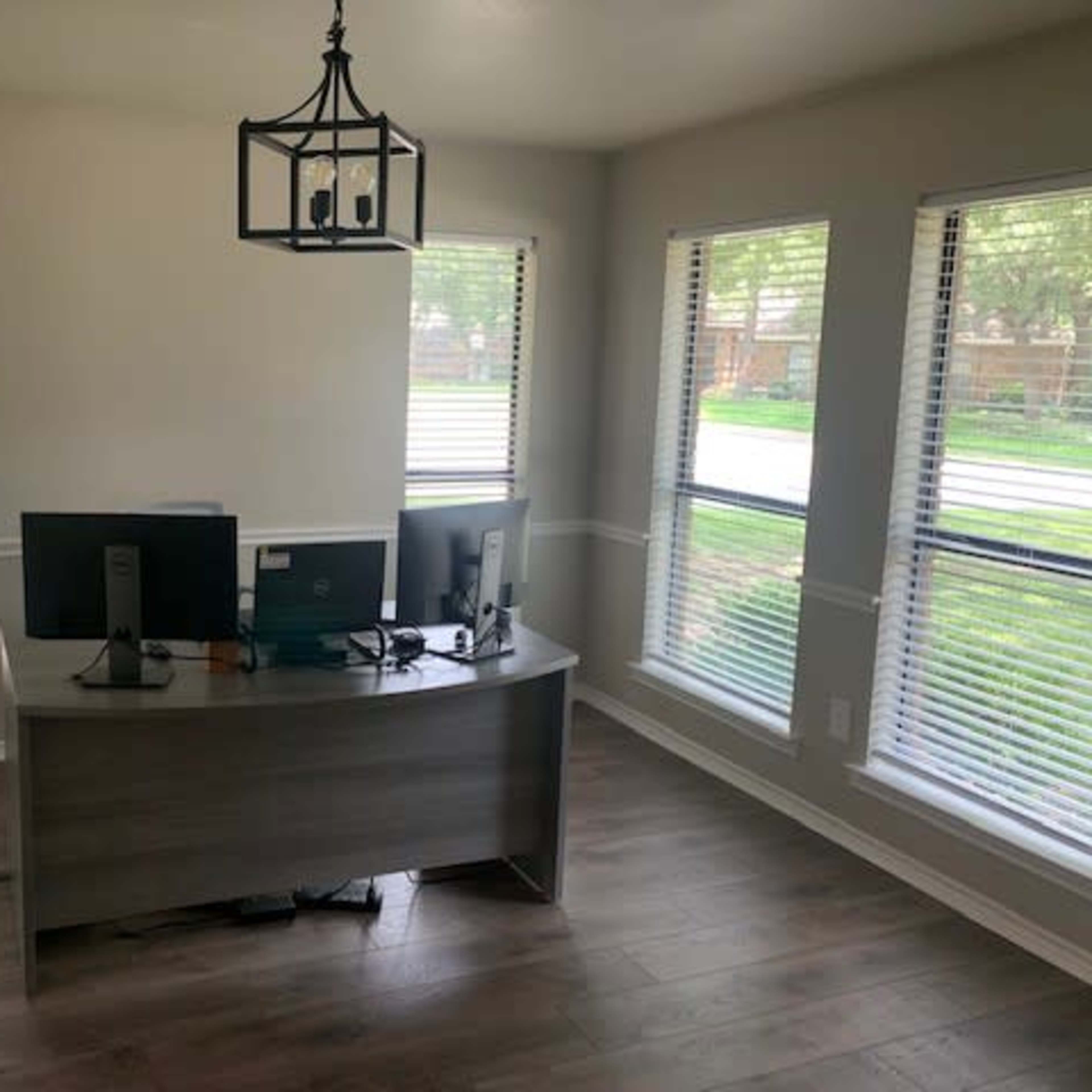 A modern office space with a wooden desk, two computer monitors, and large windows allowing natural light to enter.