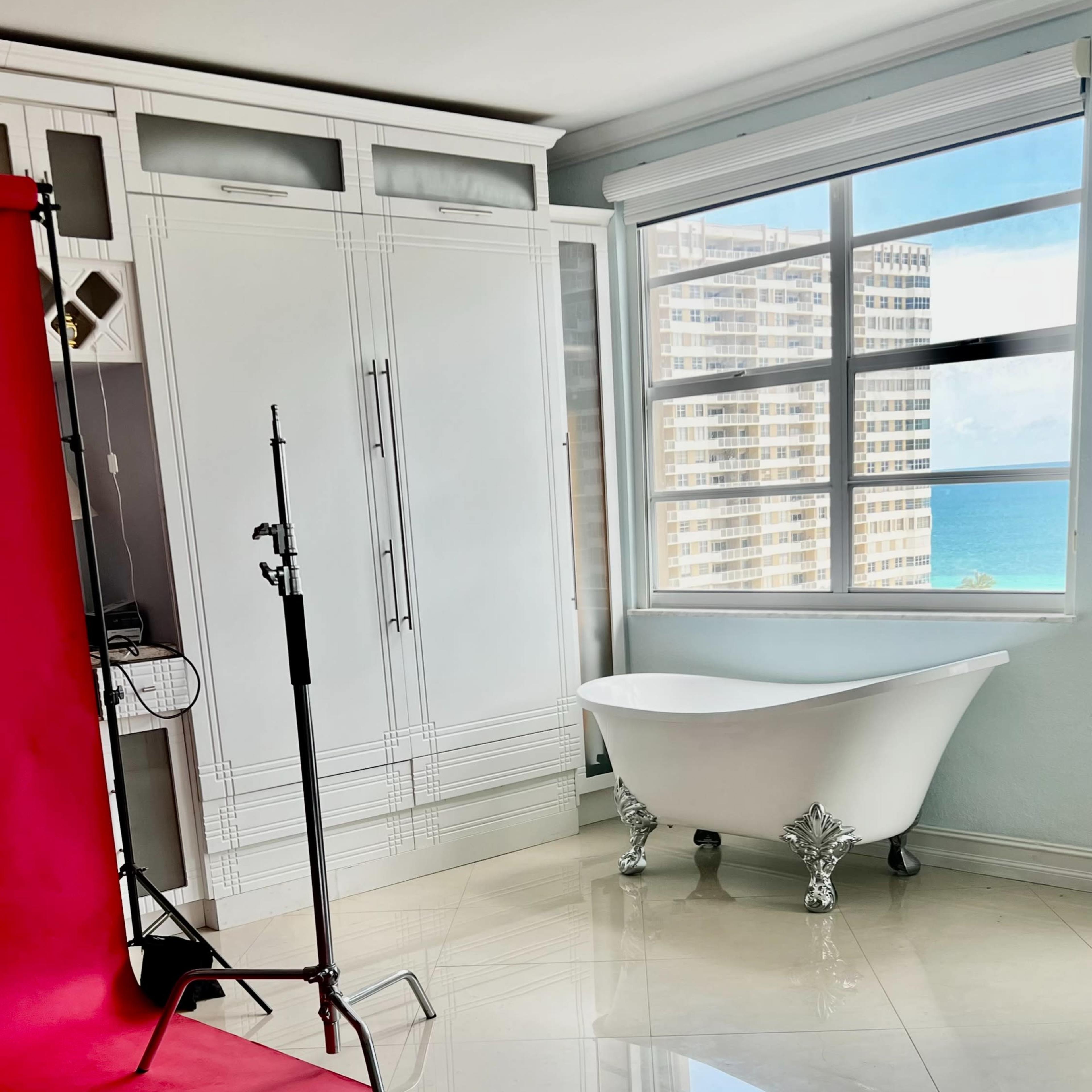 A bright interior scene with a pink backdrop, a photography stand, and a clawfoot bathtub next to a large window overlooking the ocean.