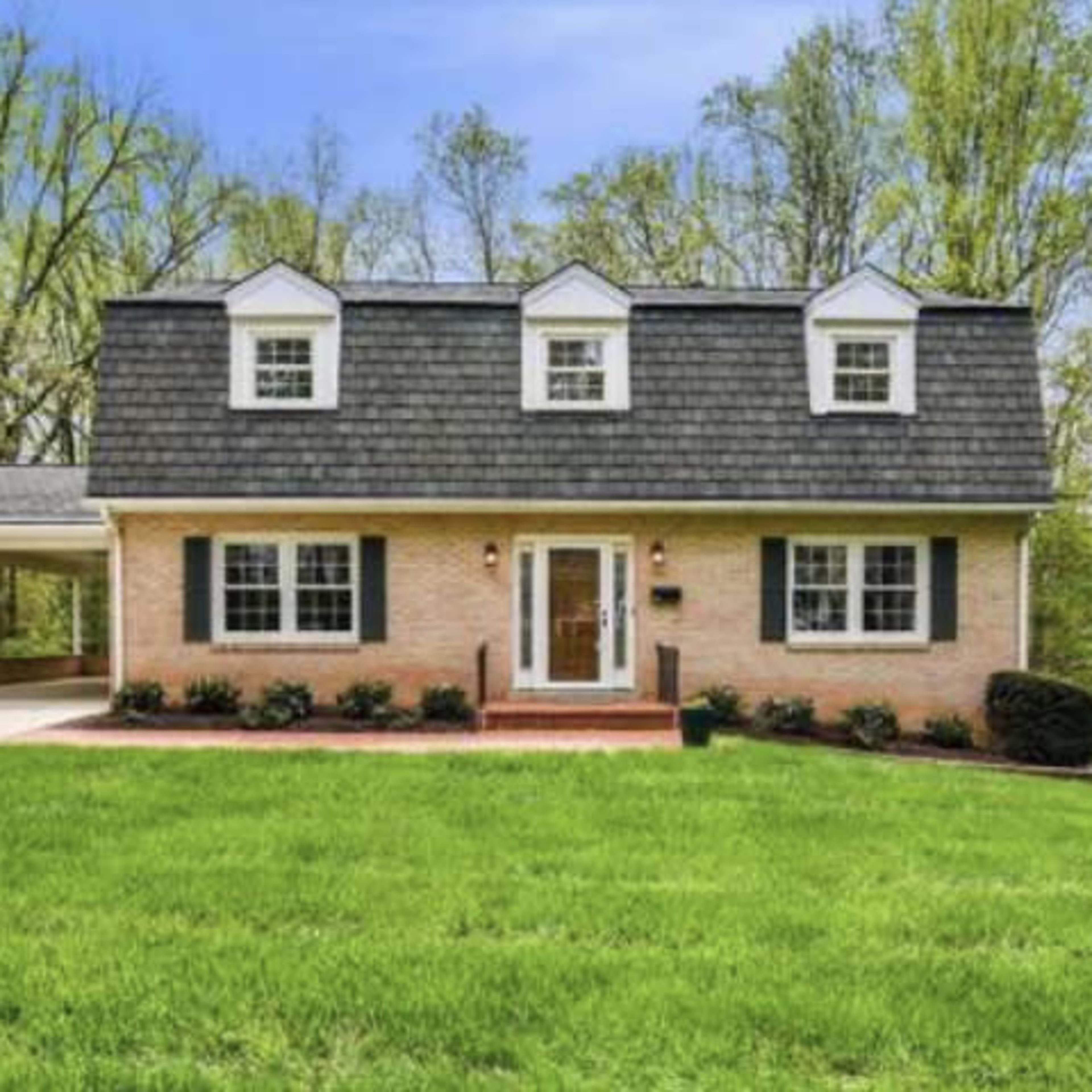 A two-story house with a gray shingles roof and a brick exterior is set on a grassy lawn surrounded by trees.