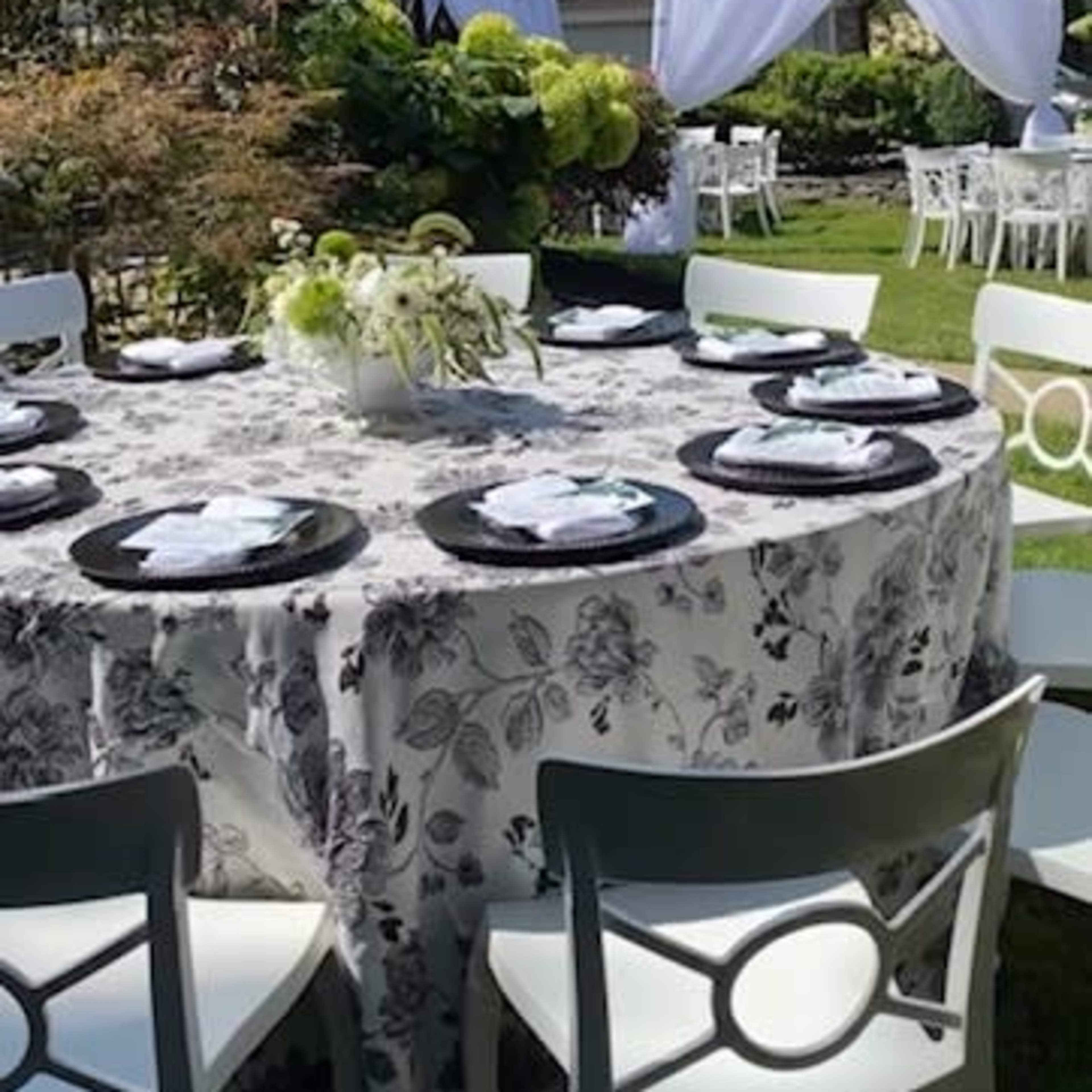 A round table set with black plates and white napkins is arranged under a blue sky in a garden, surrounded by greenery and additional seating.