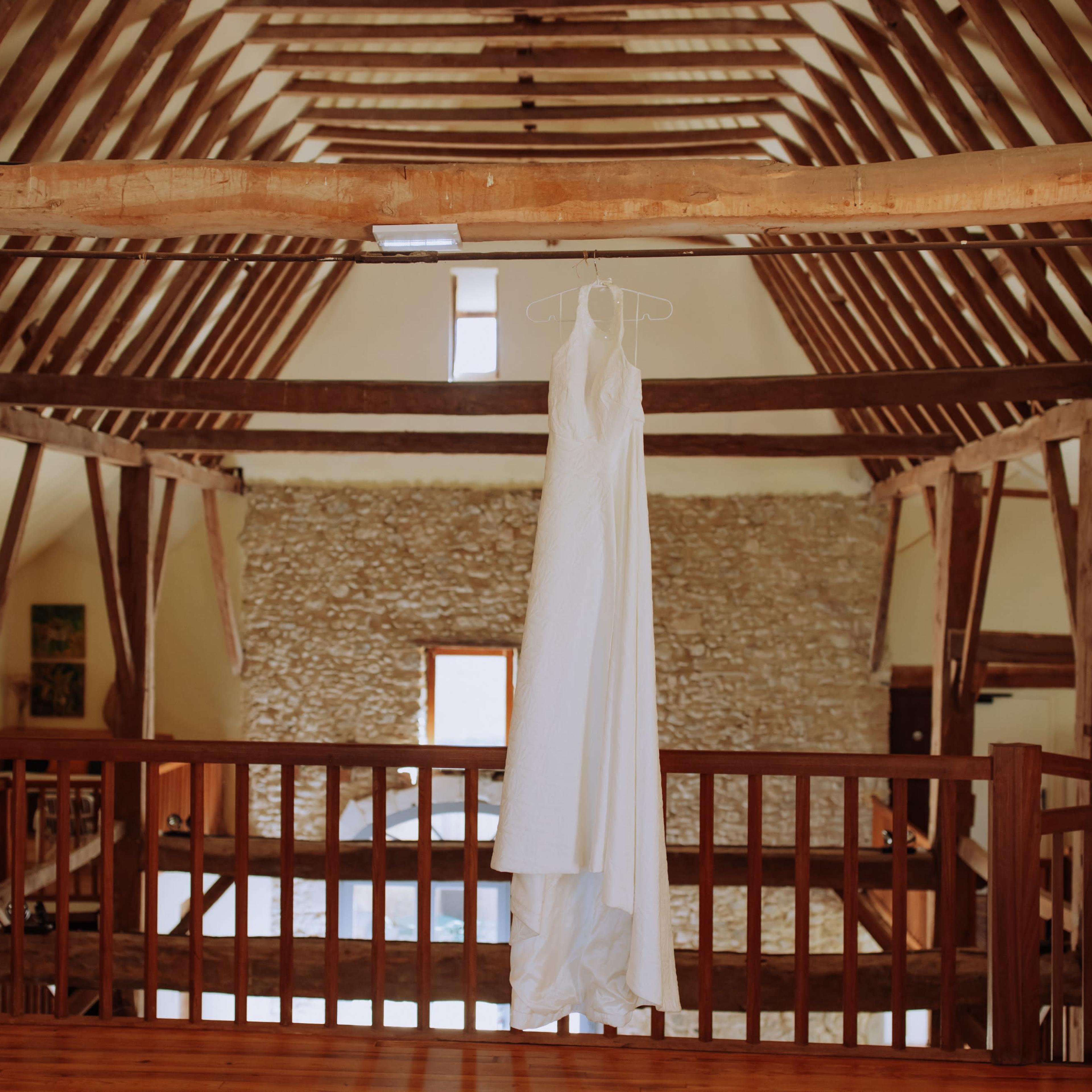 A white wedding dress is hanging on a wooden railing in a spacious, rustic room with exposed beams and stone walls.