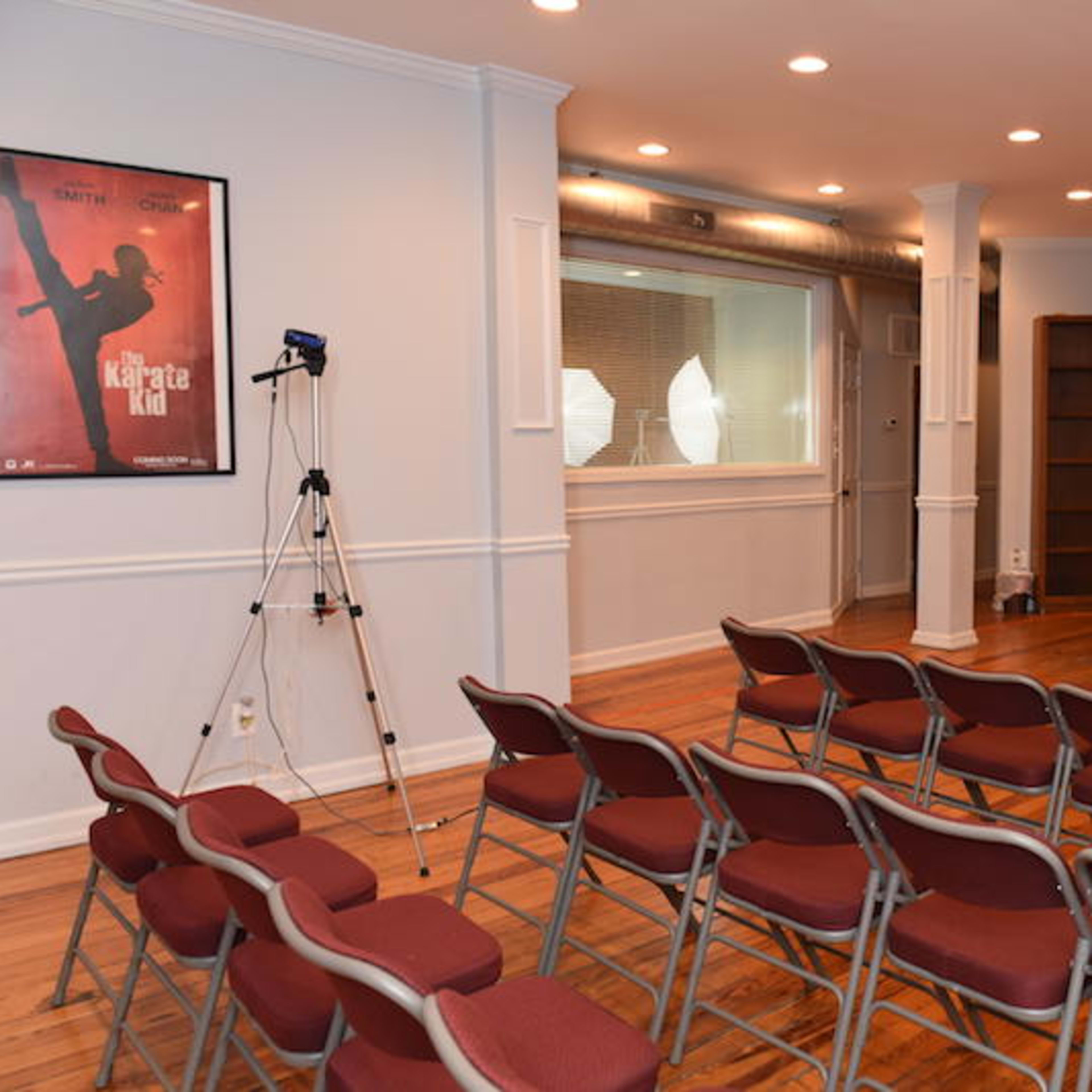 A room with wooden floors featuring rows of maroon chairs arranged for an audience, a bookshelf, and a recording setup with a camera facing a glass partition.