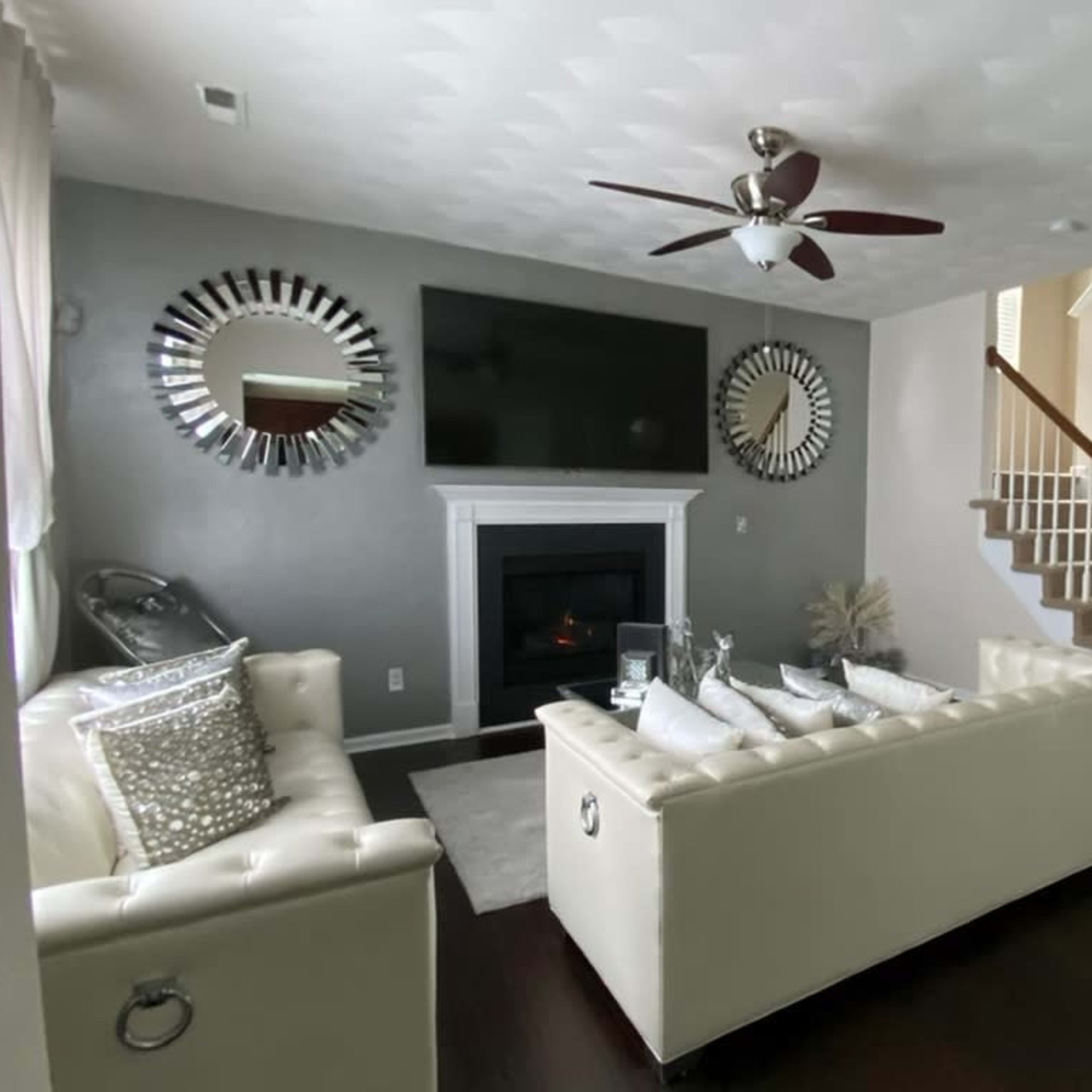 A modern living room features a white tufted sofa, a large flat-screen TV above a fireplace, and a staircase leading to an upper level.