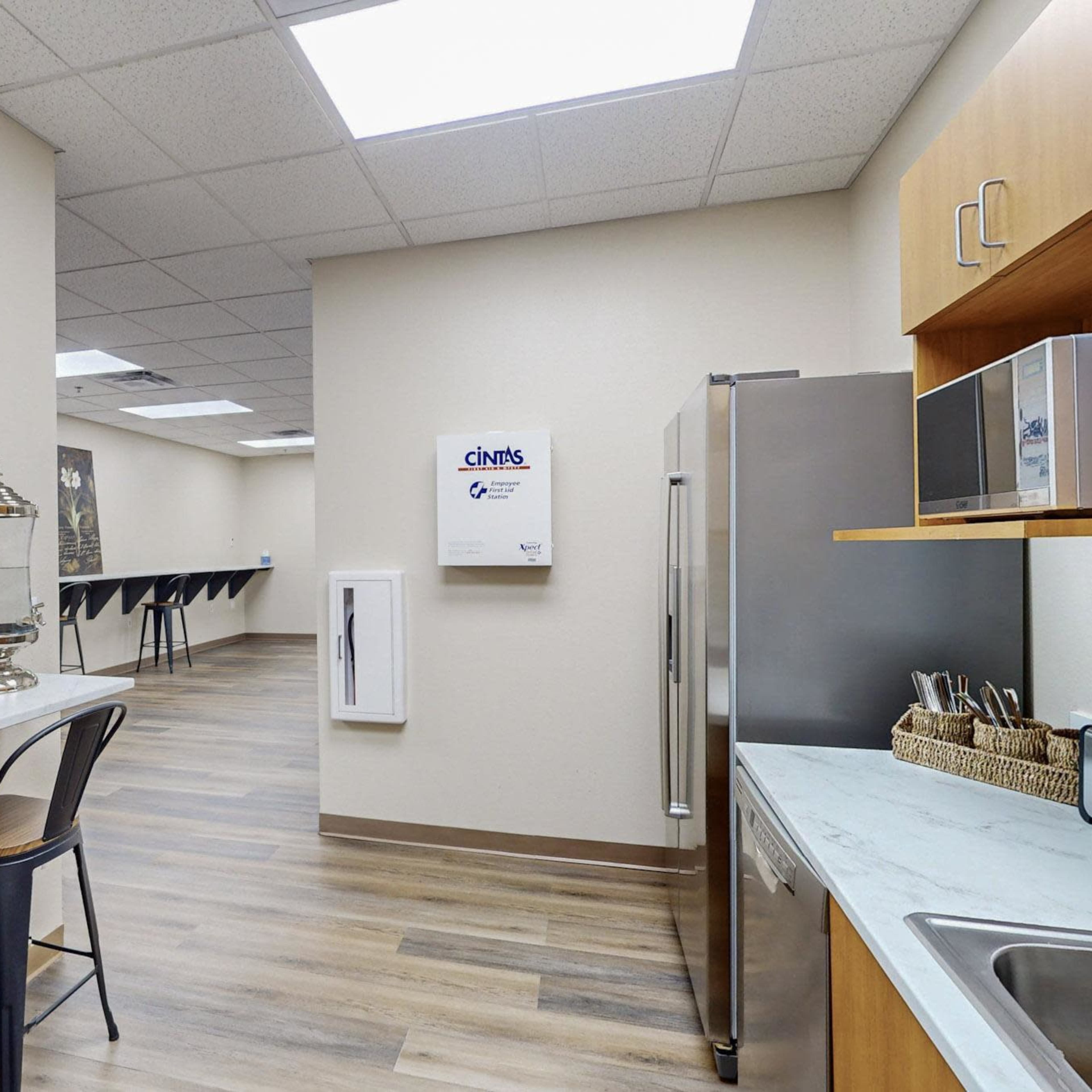 A modern kitchenette area with a counter, chairs, and stainless steel appliances, leading into a larger room with tables in the background.
