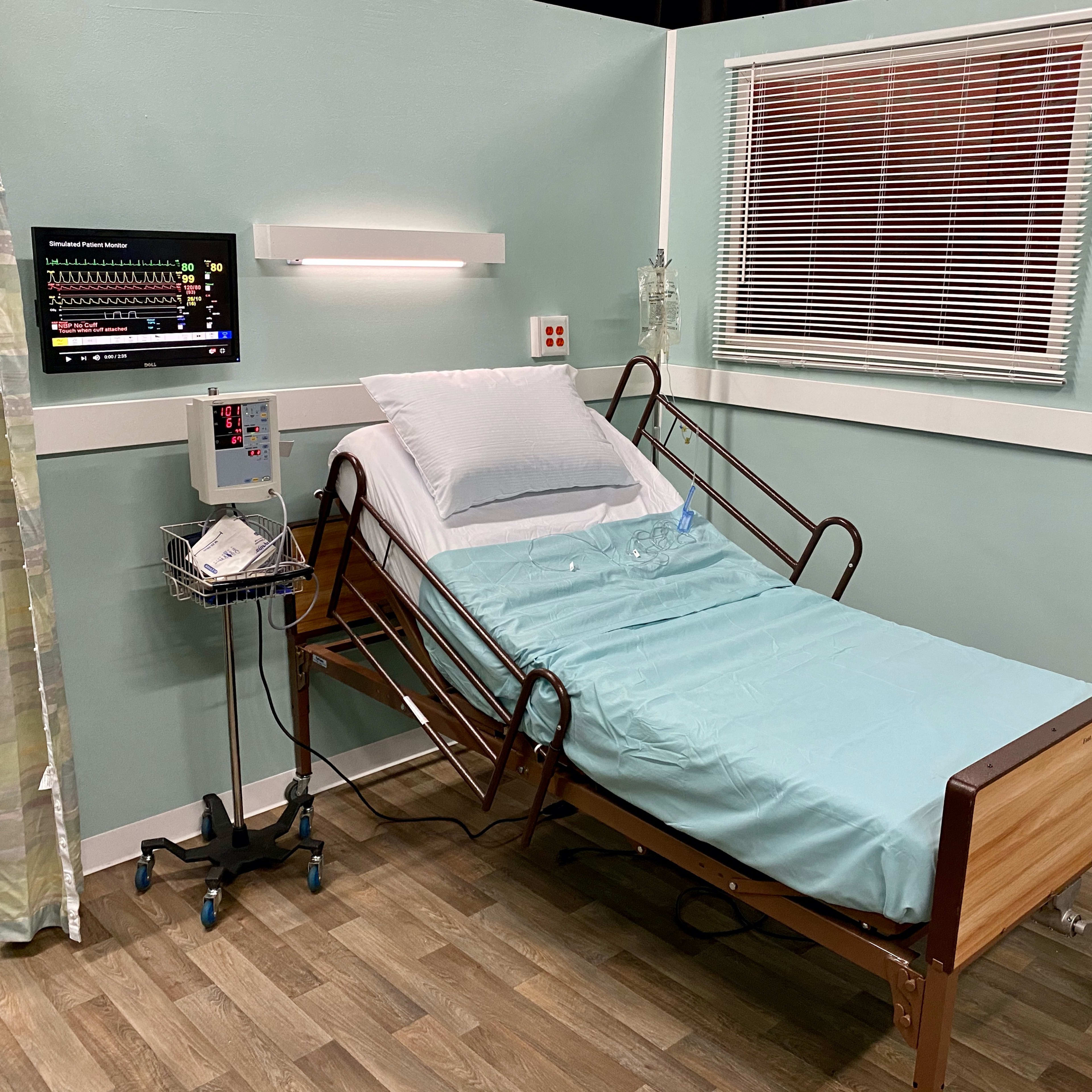 A hospital room featuring an adjustable bed with a light blue sheet, a monitor displaying medical data, and a small rolling cart with medical supplies.