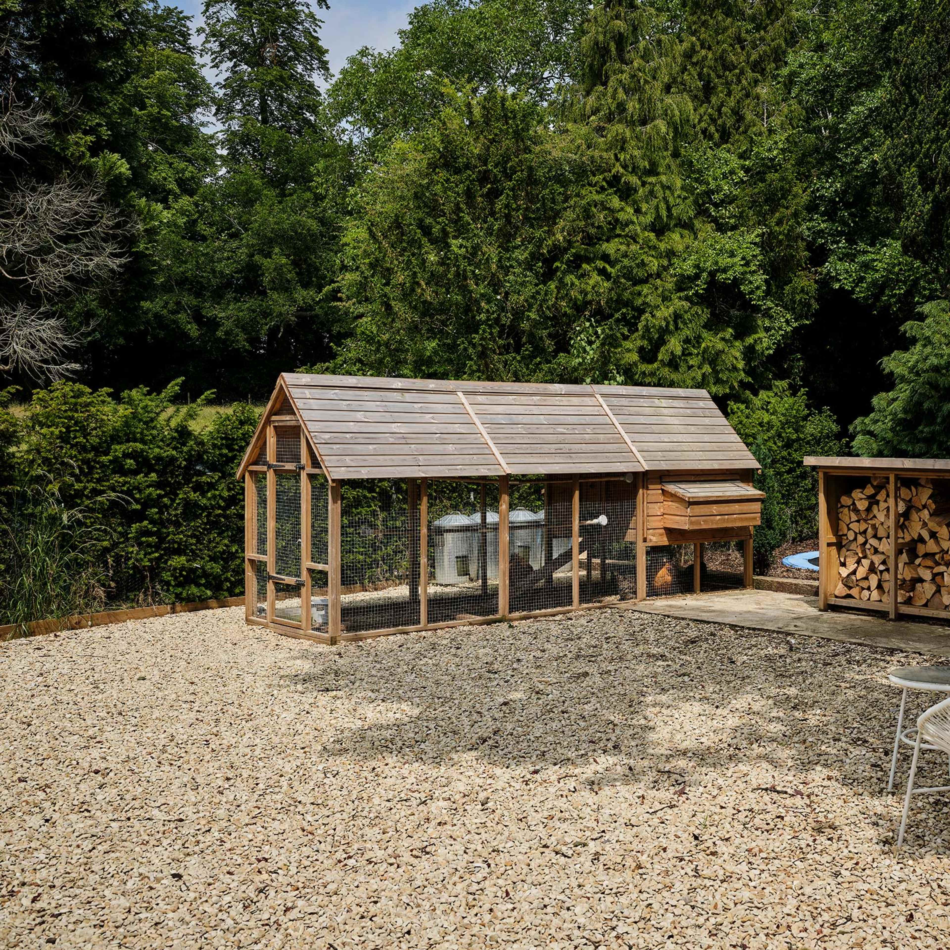 A greenhouse with a chicken coop beside a woodpile, situated on a gravel area surrounded by trees.