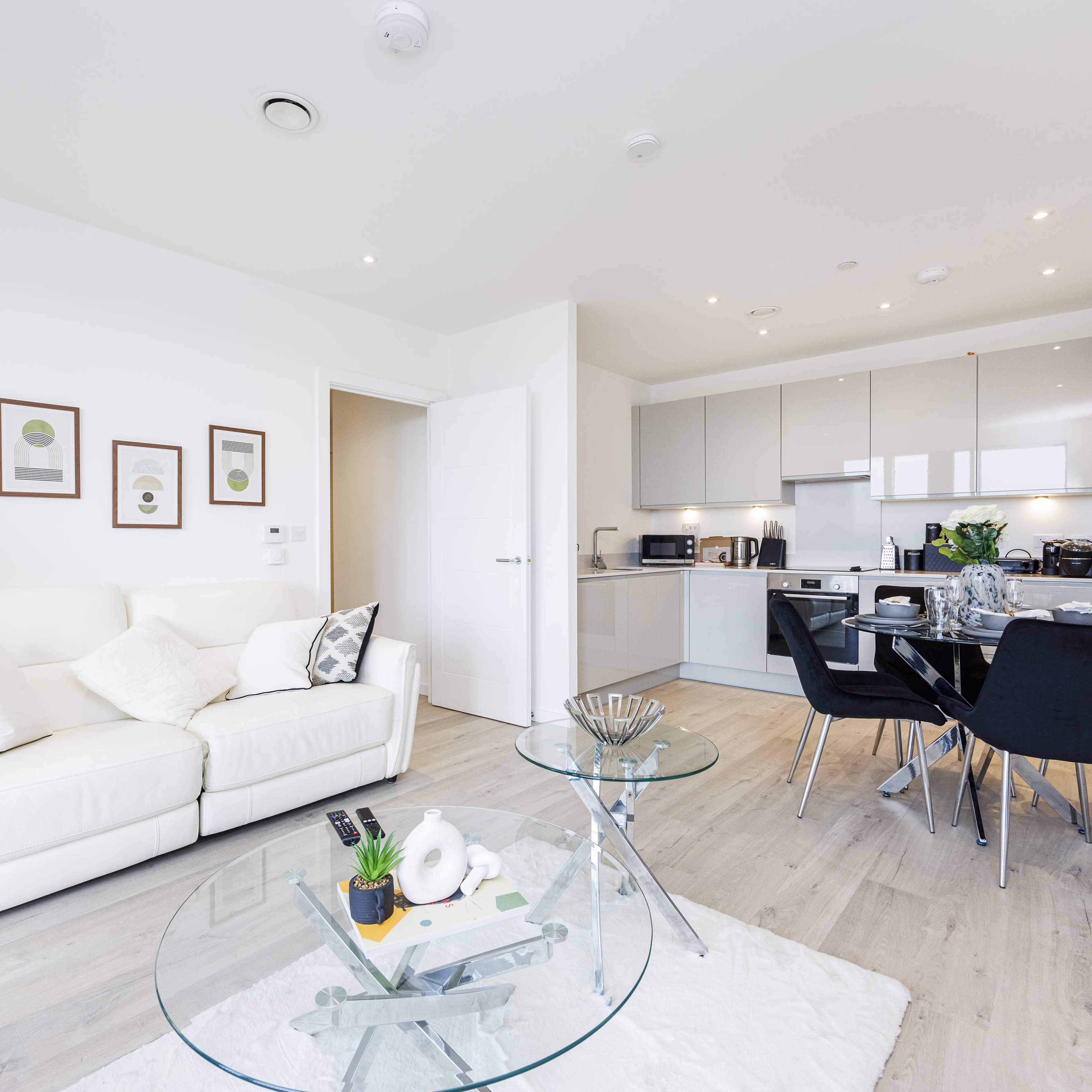 A modern living room with a white sofa and a glass coffee table, leading into a sleek kitchen with gray cabinets and stainless steel appliances.