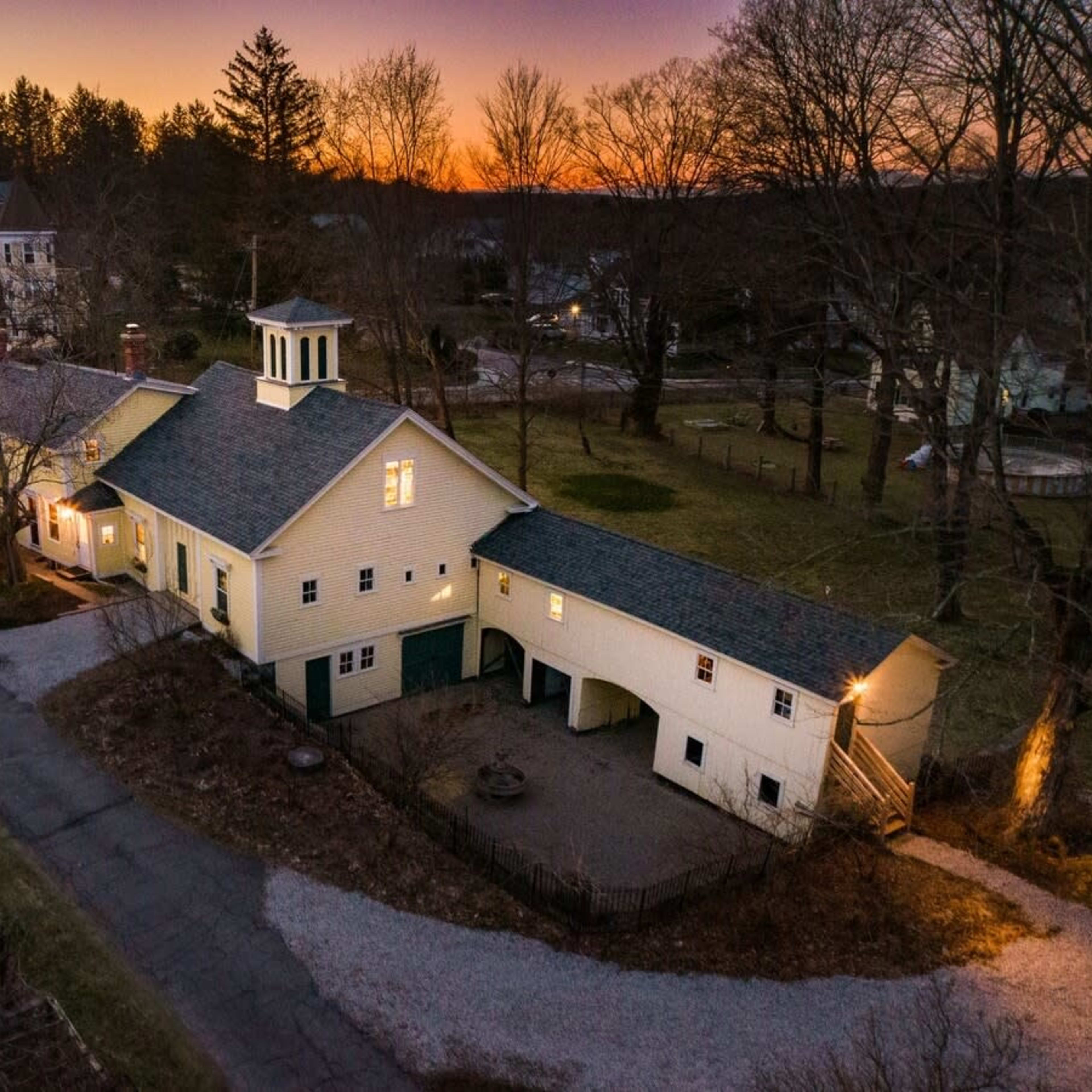 A large, two-story yellow house with a cupola is situated at the end of a gravel driveway, surrounded by trees during sunset.
