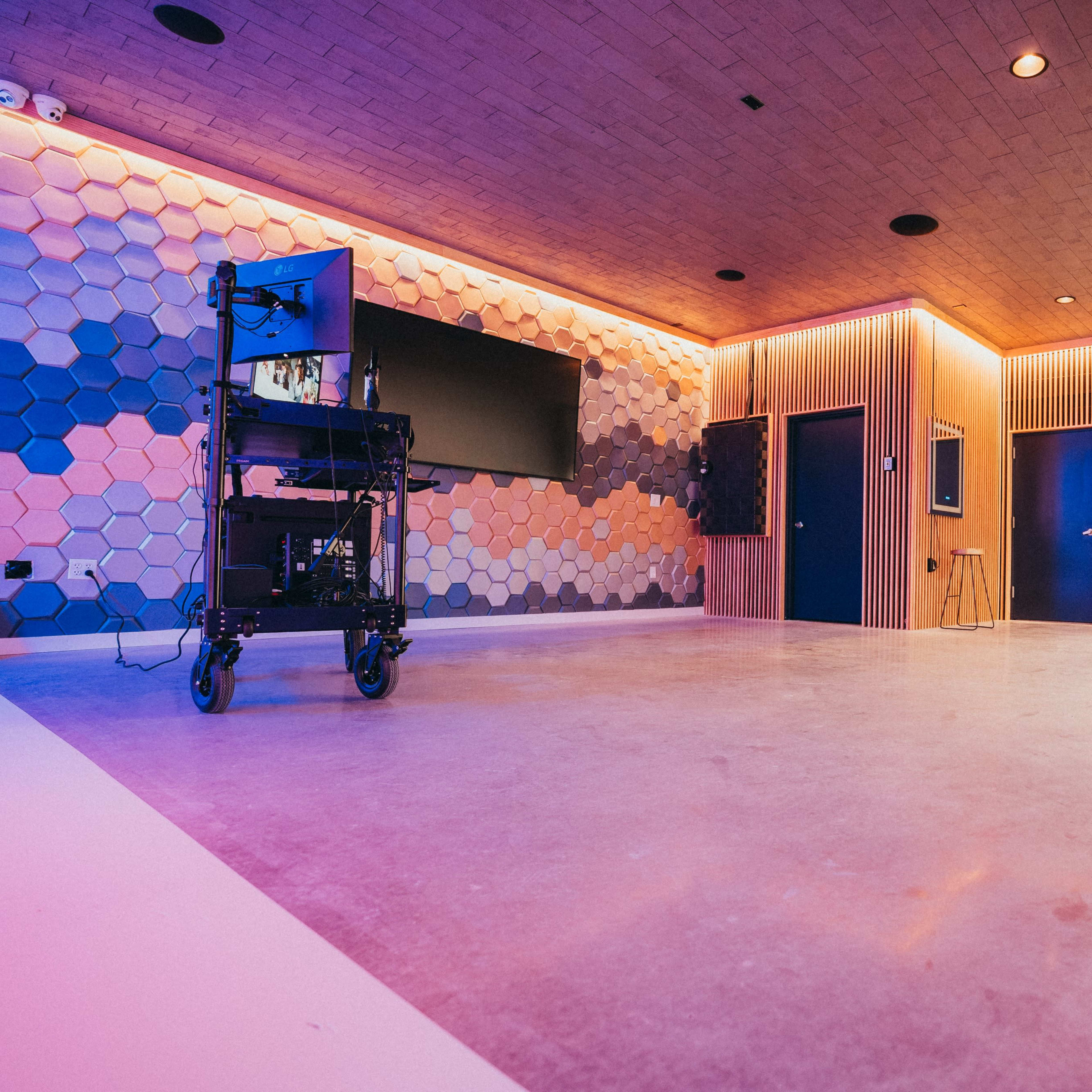 A modern, empty room with hexagonal wall patterns and ambient lighting, featuring a television mounted on one wall and a mobile cart in the center.