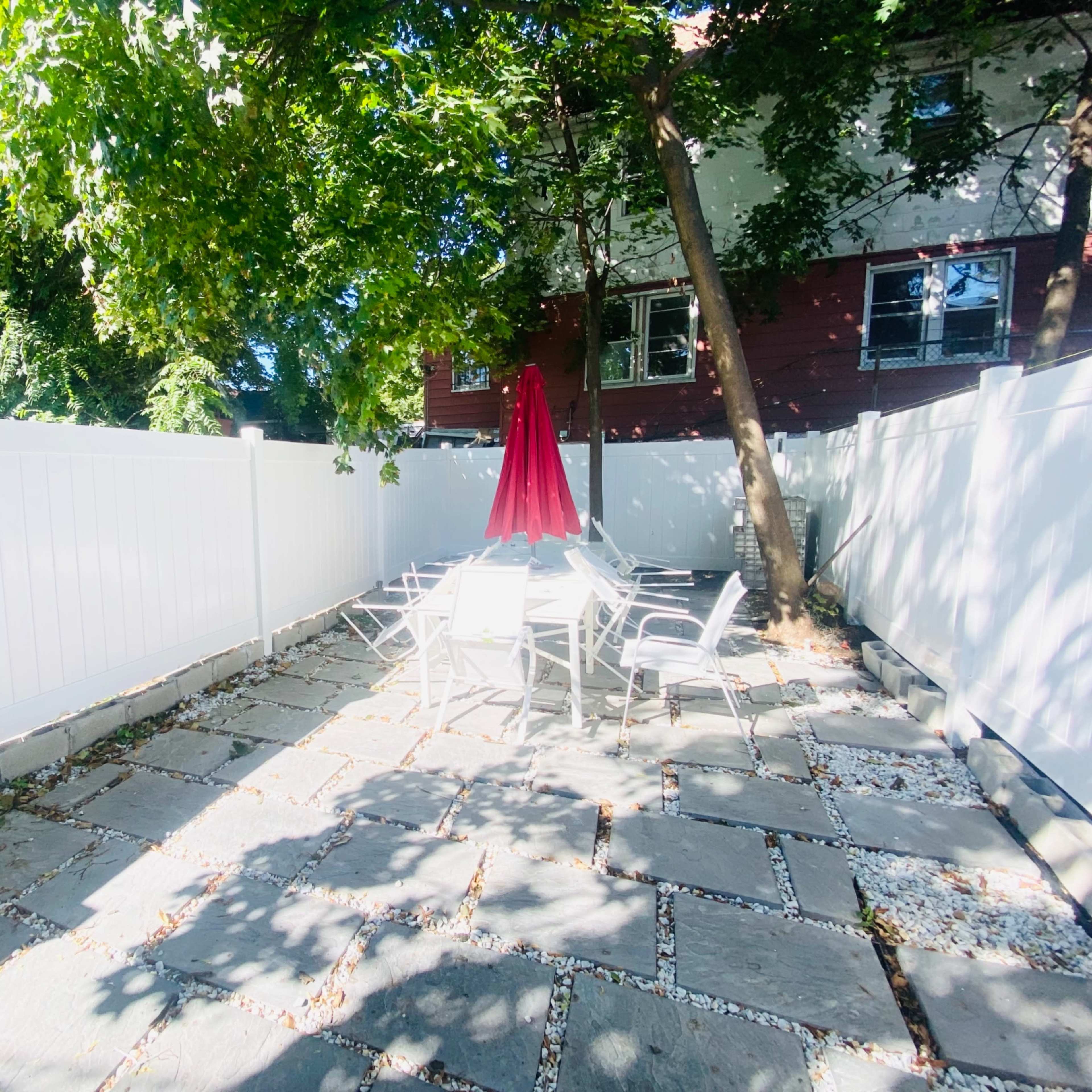 A fenced patio area features a table and chairs shaded by a red umbrella, surrounded by trees and a white fence.