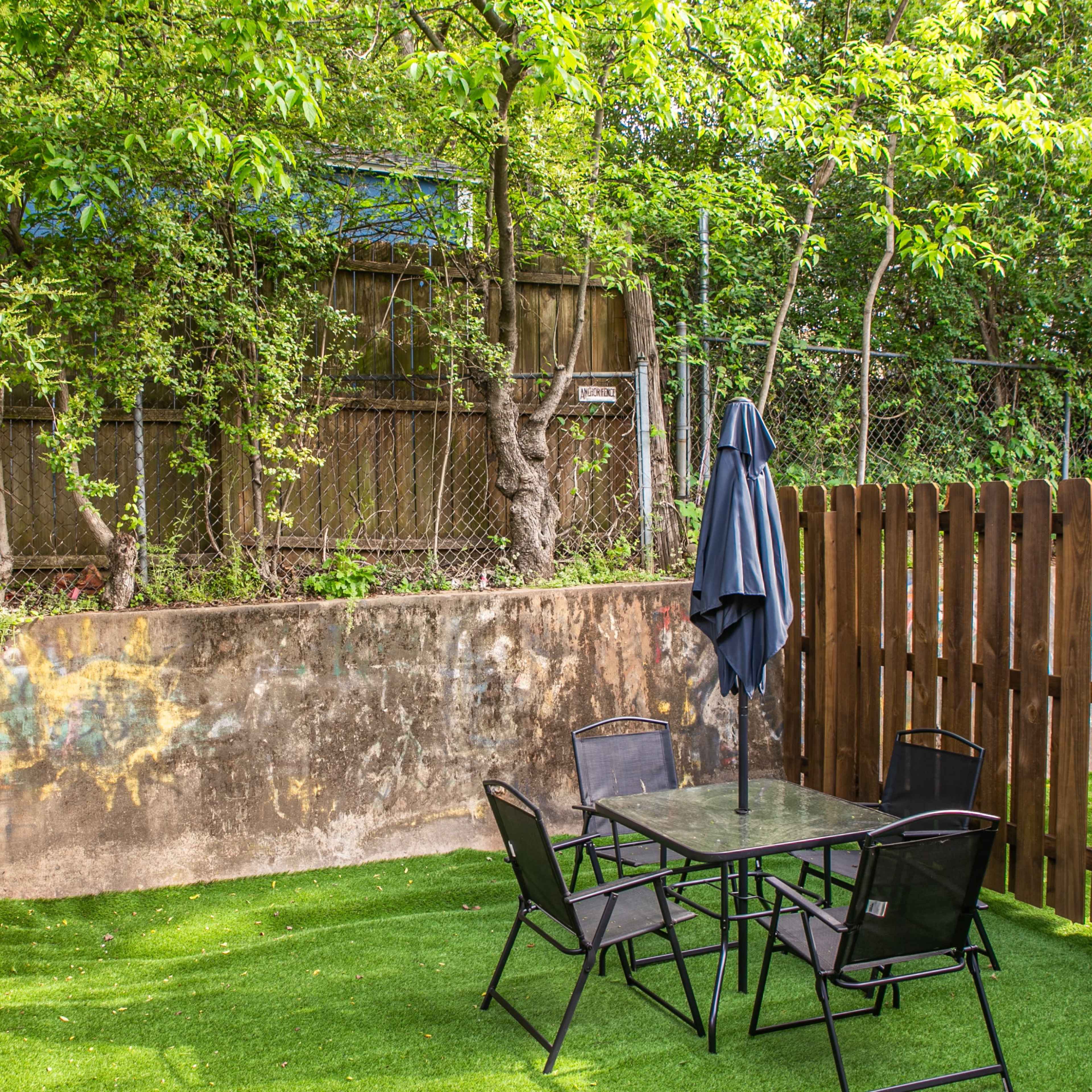 A small enclosed backyard features a glass table surrounded by four black chairs, with artificial grass and a wooden fence.
