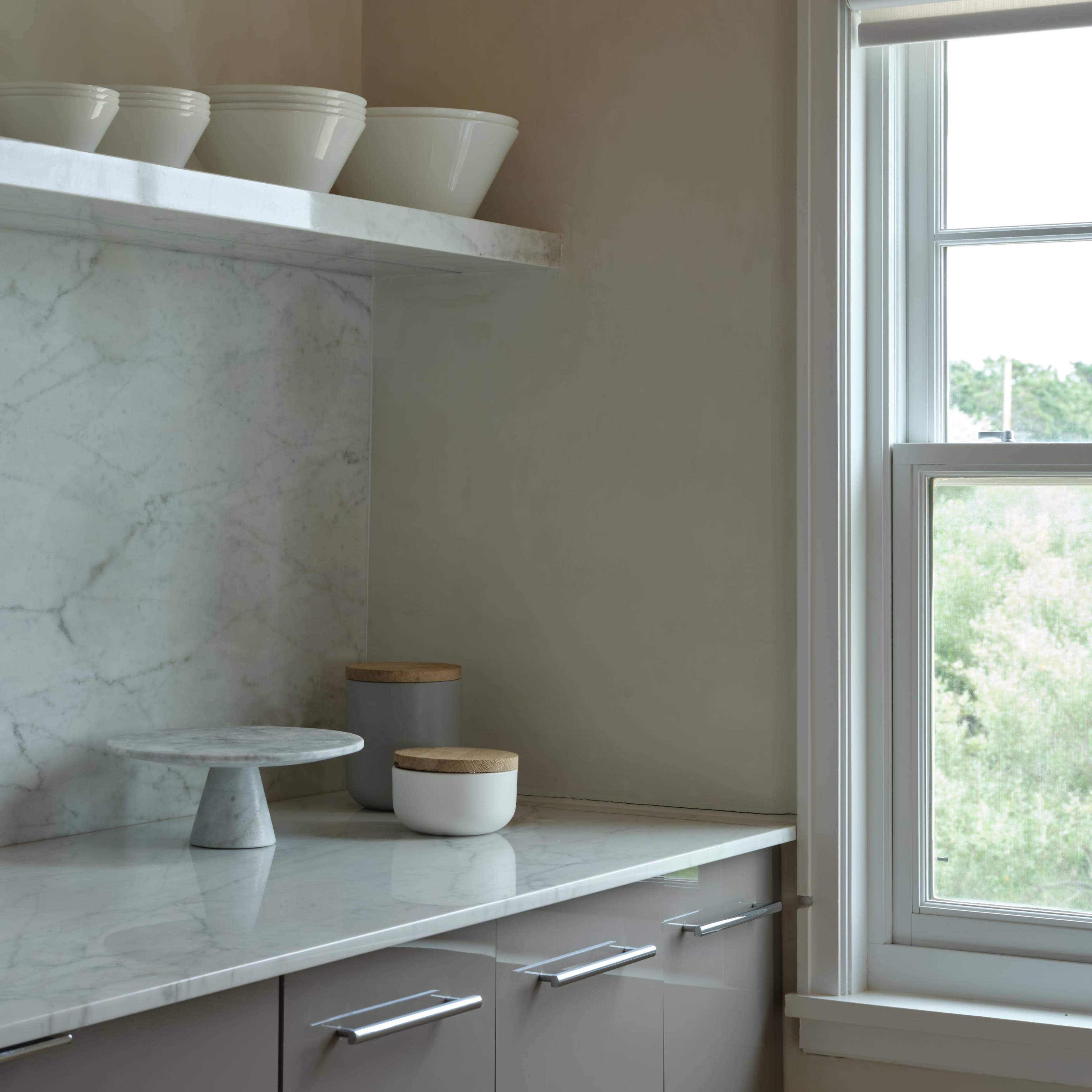 A modern kitchen corner with a marble countertop, a cake stand, and white bowls on a shelf above, beside a window with a view of greenery.