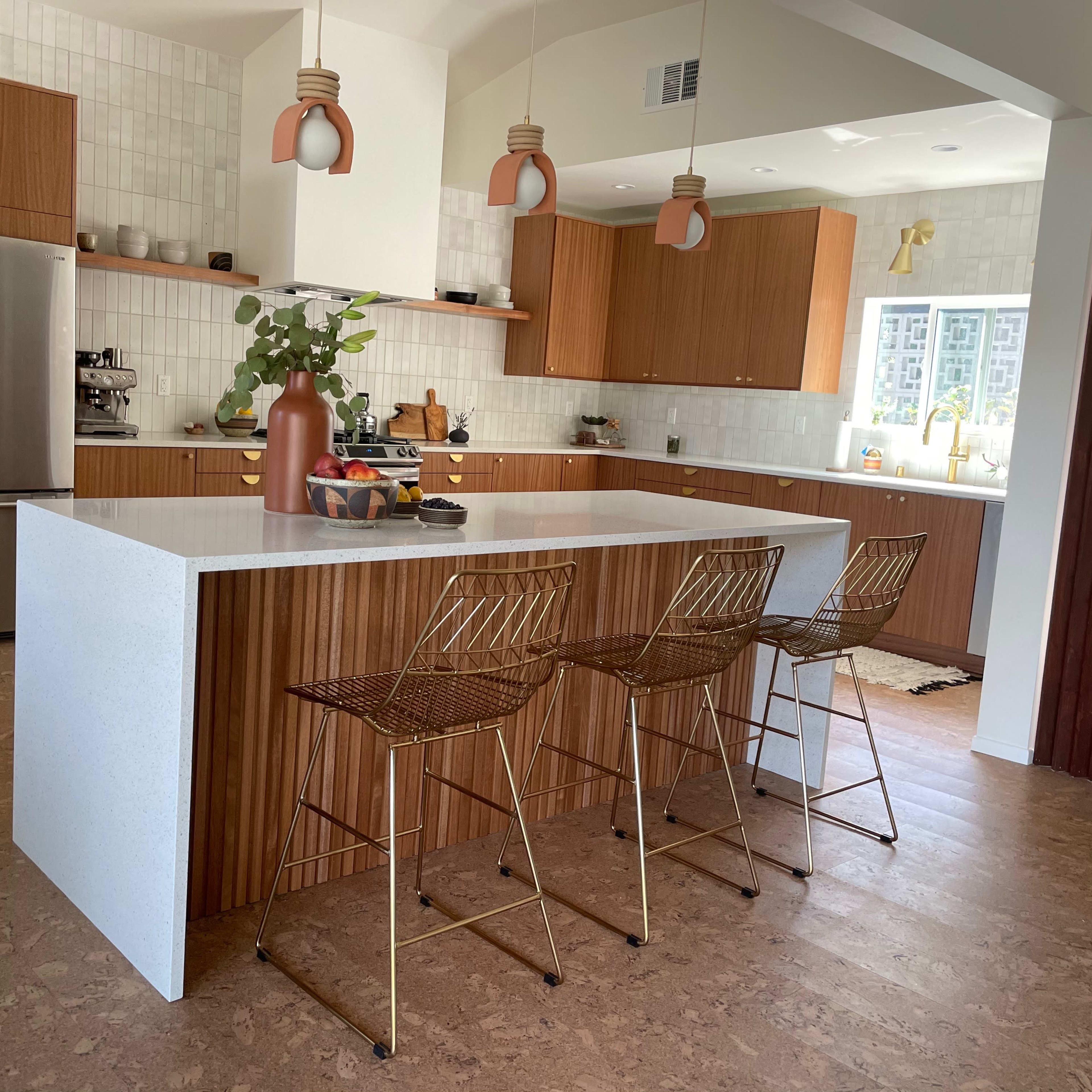 A modern kitchen features a large white island with three metal bar stools, wooden cabinetry, and light fixtures hanging above.
