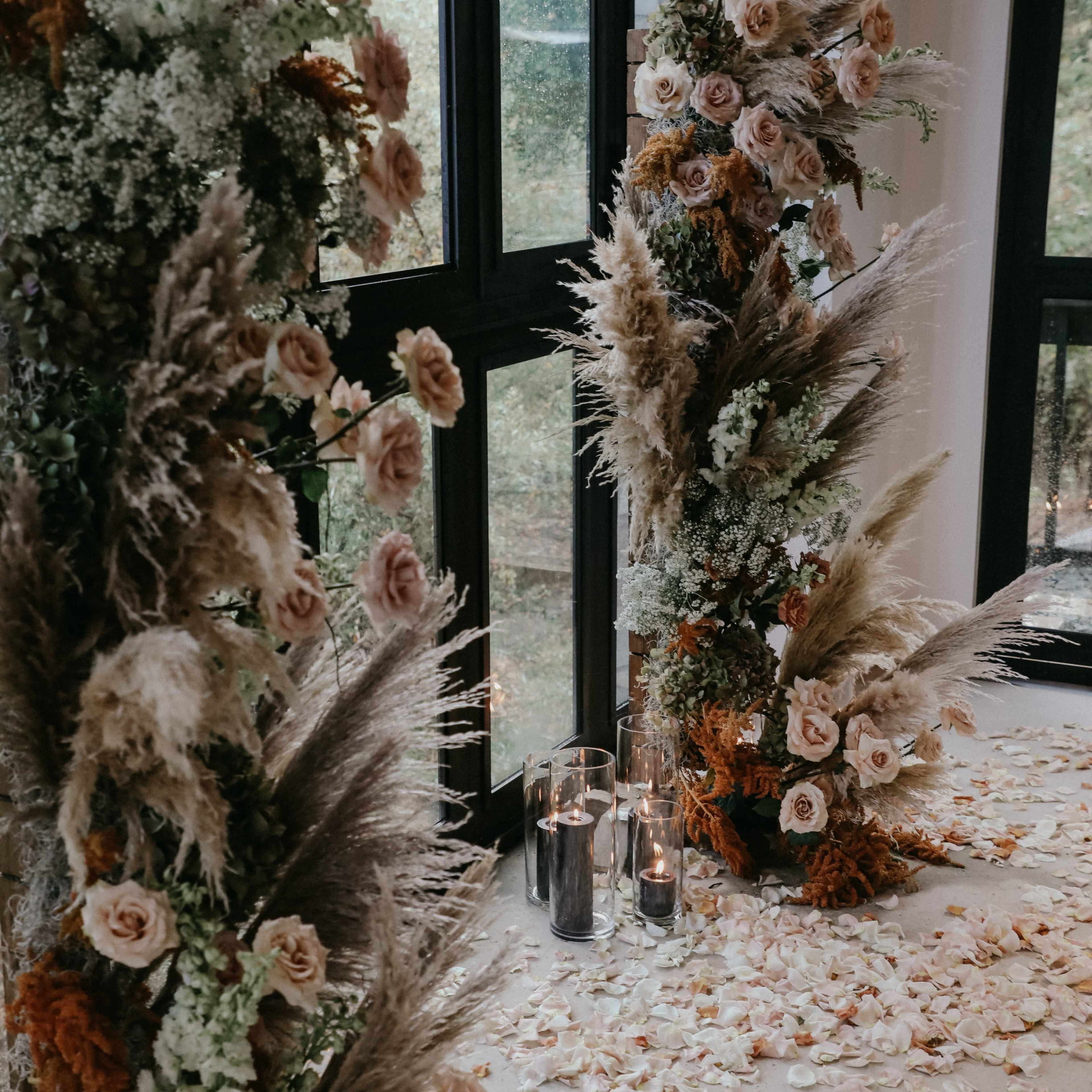 Two floral arrangements made of roses and pampas grass beside a carpet of rose petals and lit candles on the floor.