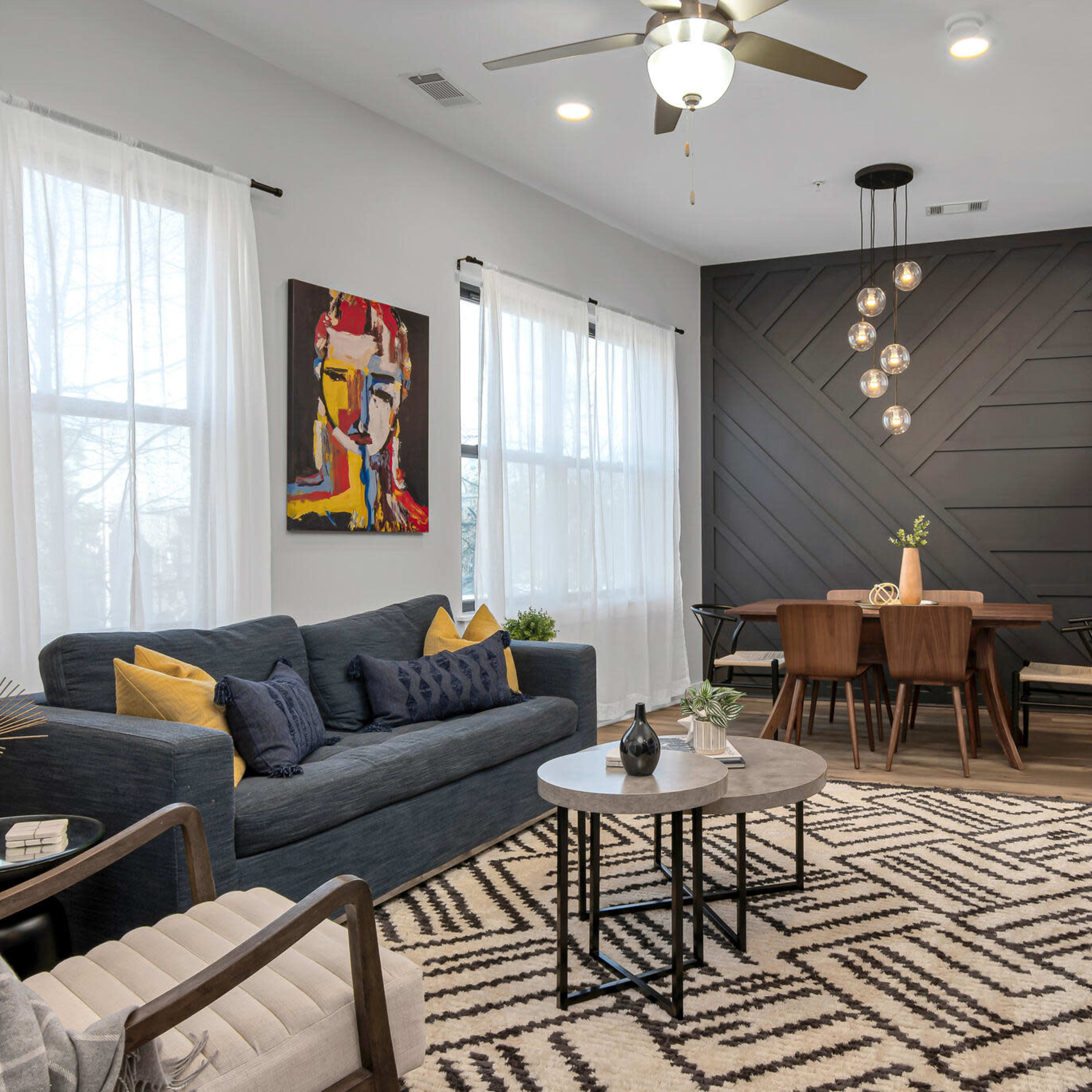 A modern living room with a blue sofa, a round coffee table, and a dining area featuring a wooden table and chairs, all set against a backdrop of large windows and stylish wall designs.