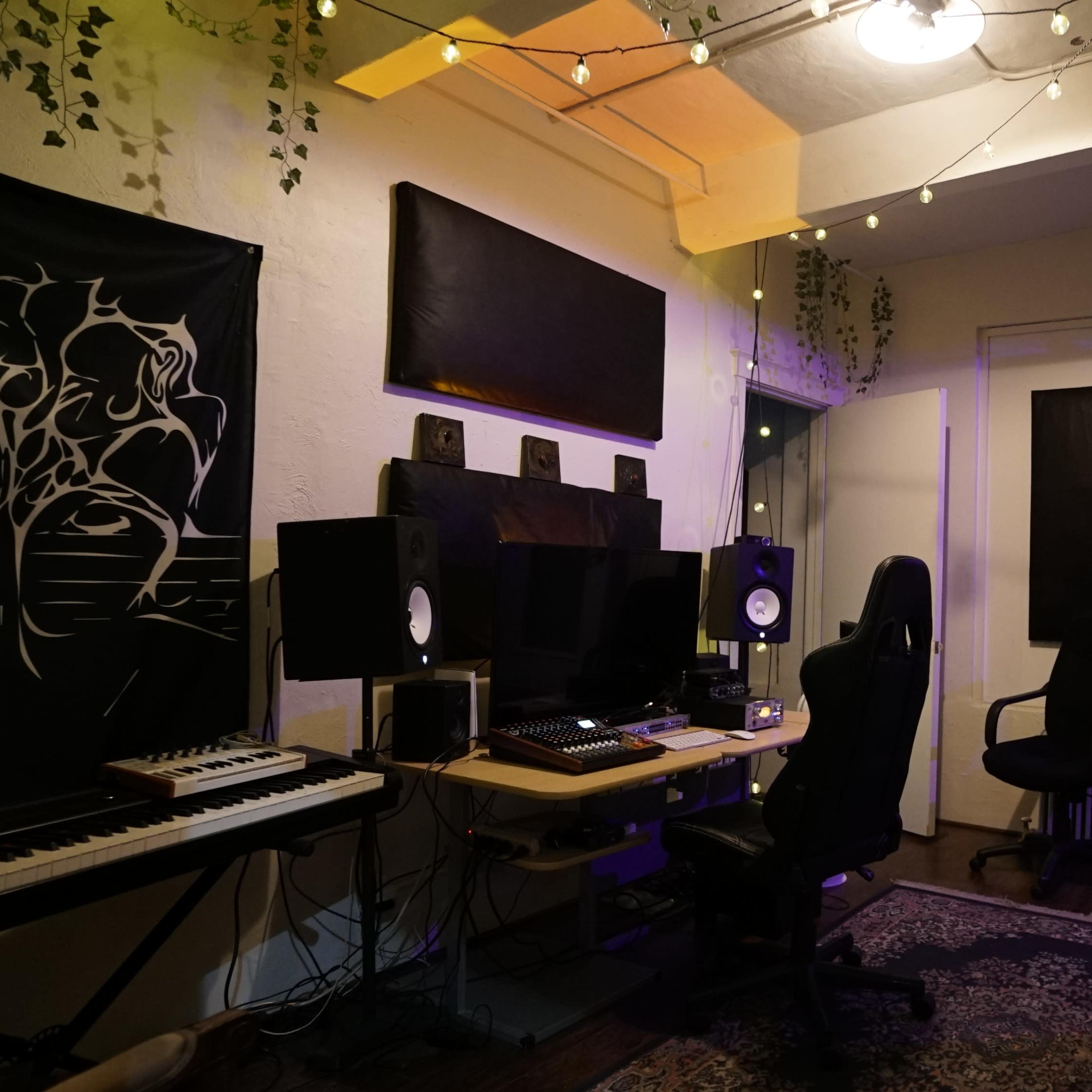 A music production studio with a keyboard, audio monitors, two chairs, and decorative plants hanging from the ceiling.