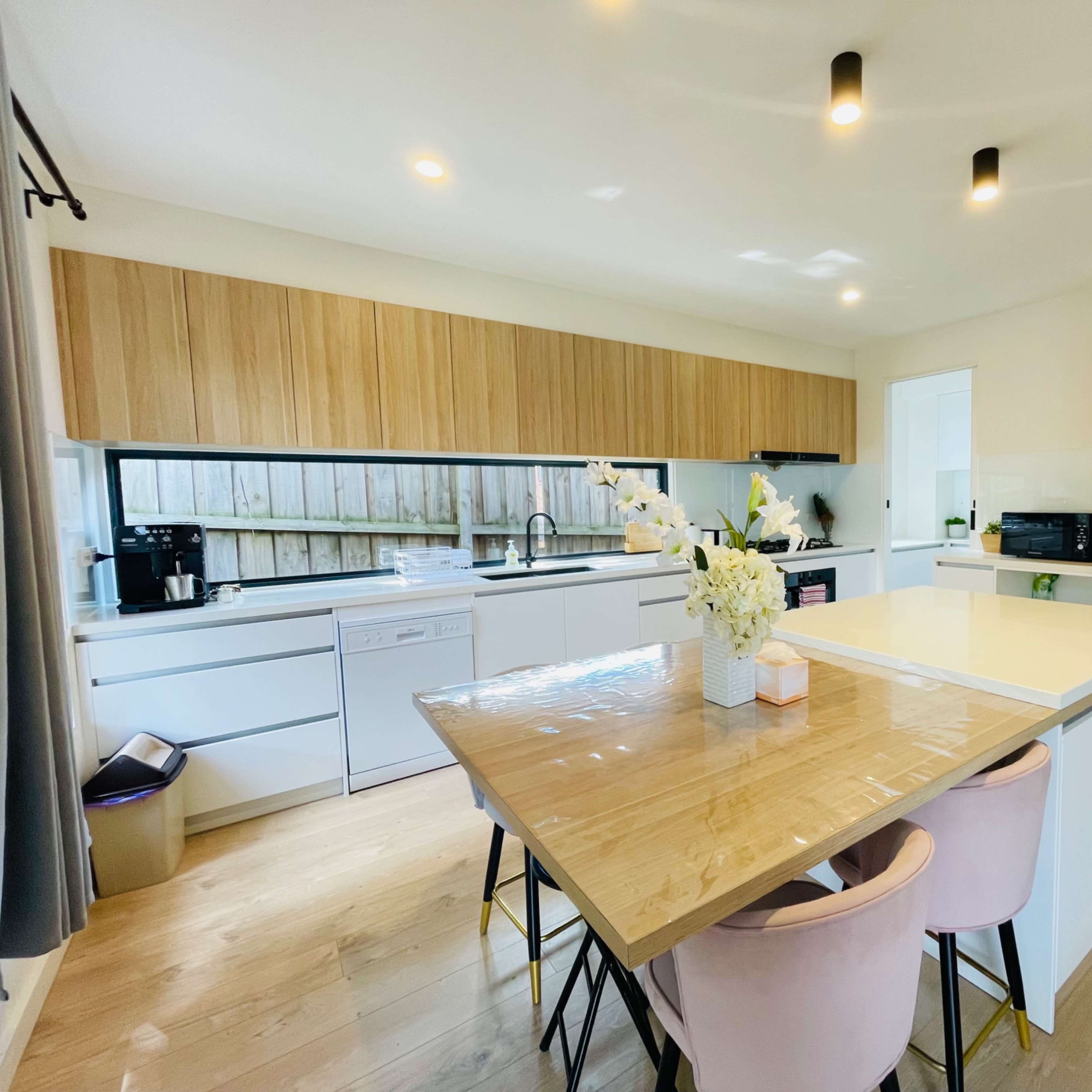 A modern kitchen features light wood cabinetry, white countertops, and a dining table with pink chairs, illuminated by overhead lighting.