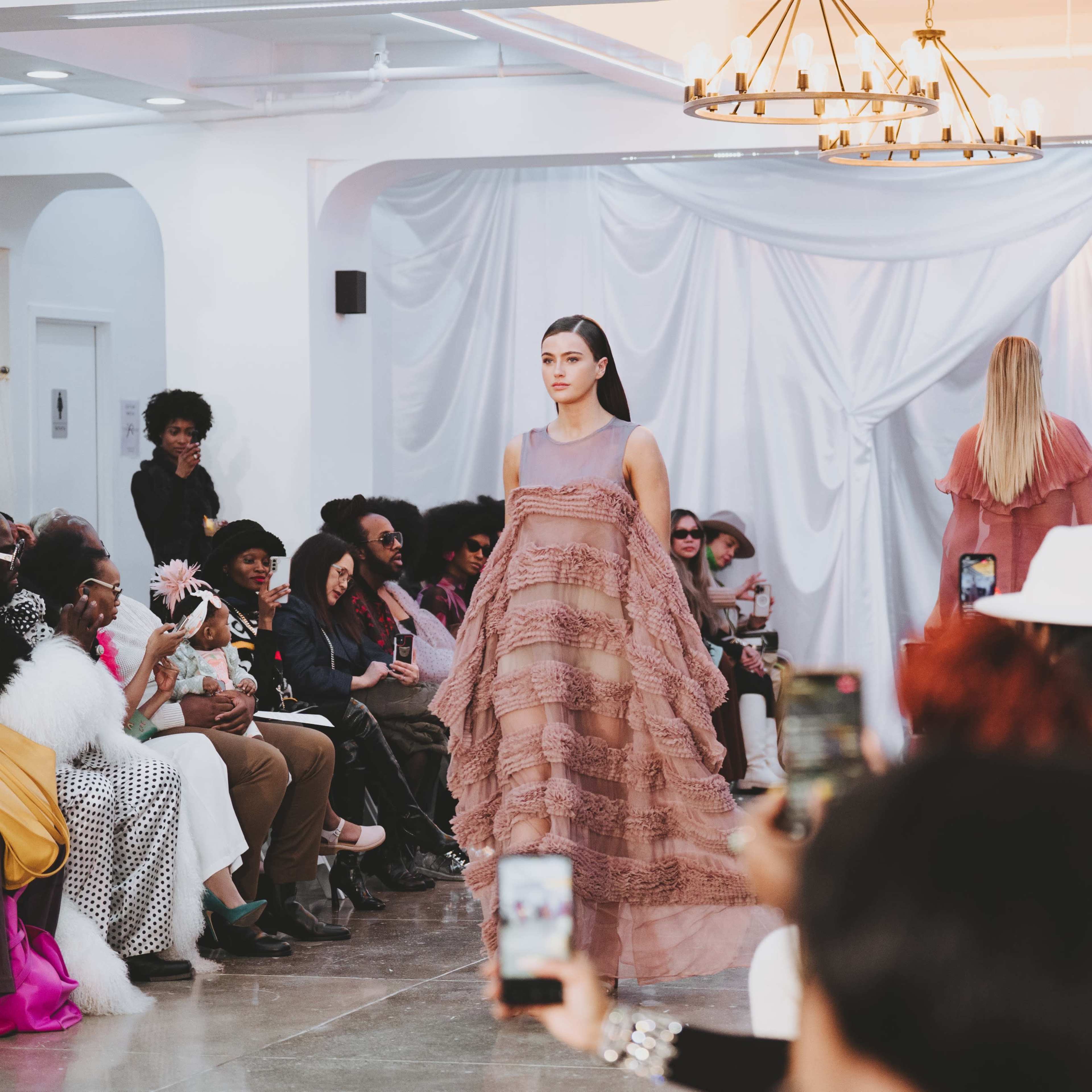 A model walks down the runway in a layered, blush-colored dress, while an audience of diverse spectators watches the fashion show.