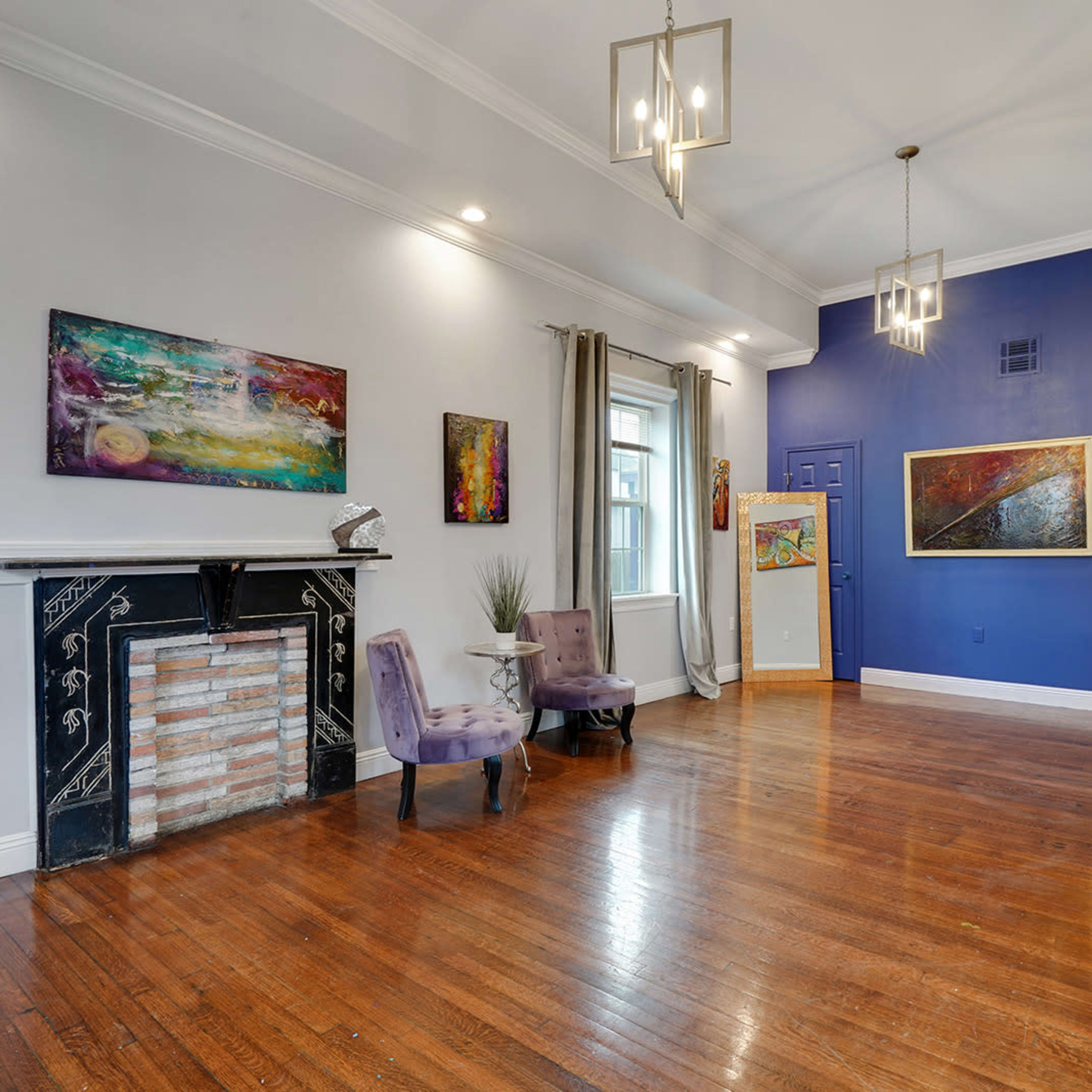 A spacious living area with polished hardwood floors, colorful artwork on the walls, and two chairs in front of a decorative fireplace.
