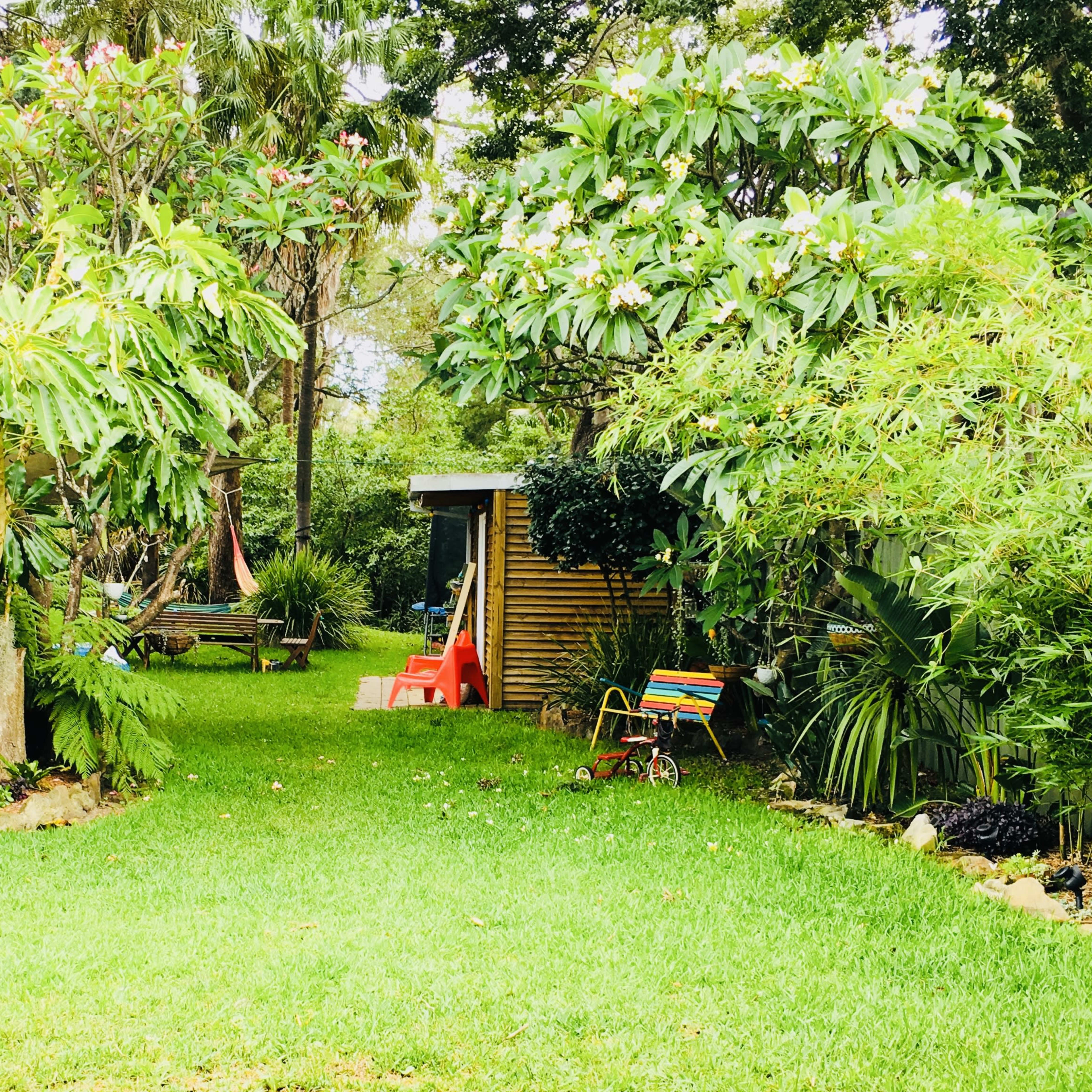 A lush garden features green grass, tropical plants, and a small shed, with a children's toy and red plastic chair visible along the pathway.
