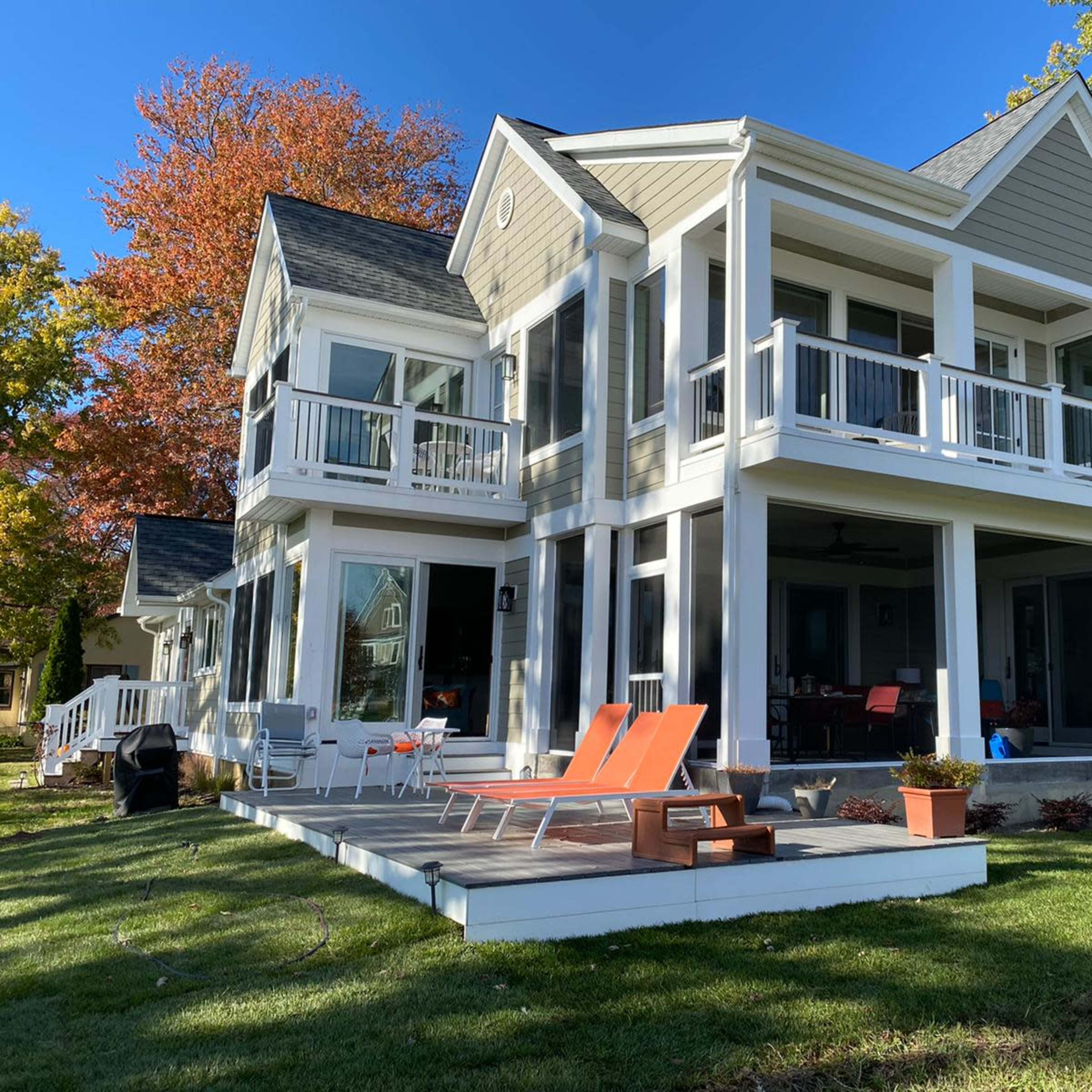 A modern two-story house with large windows, a deck, and orange lounge chairs in a grassy area, framed by trees displaying fall colors.