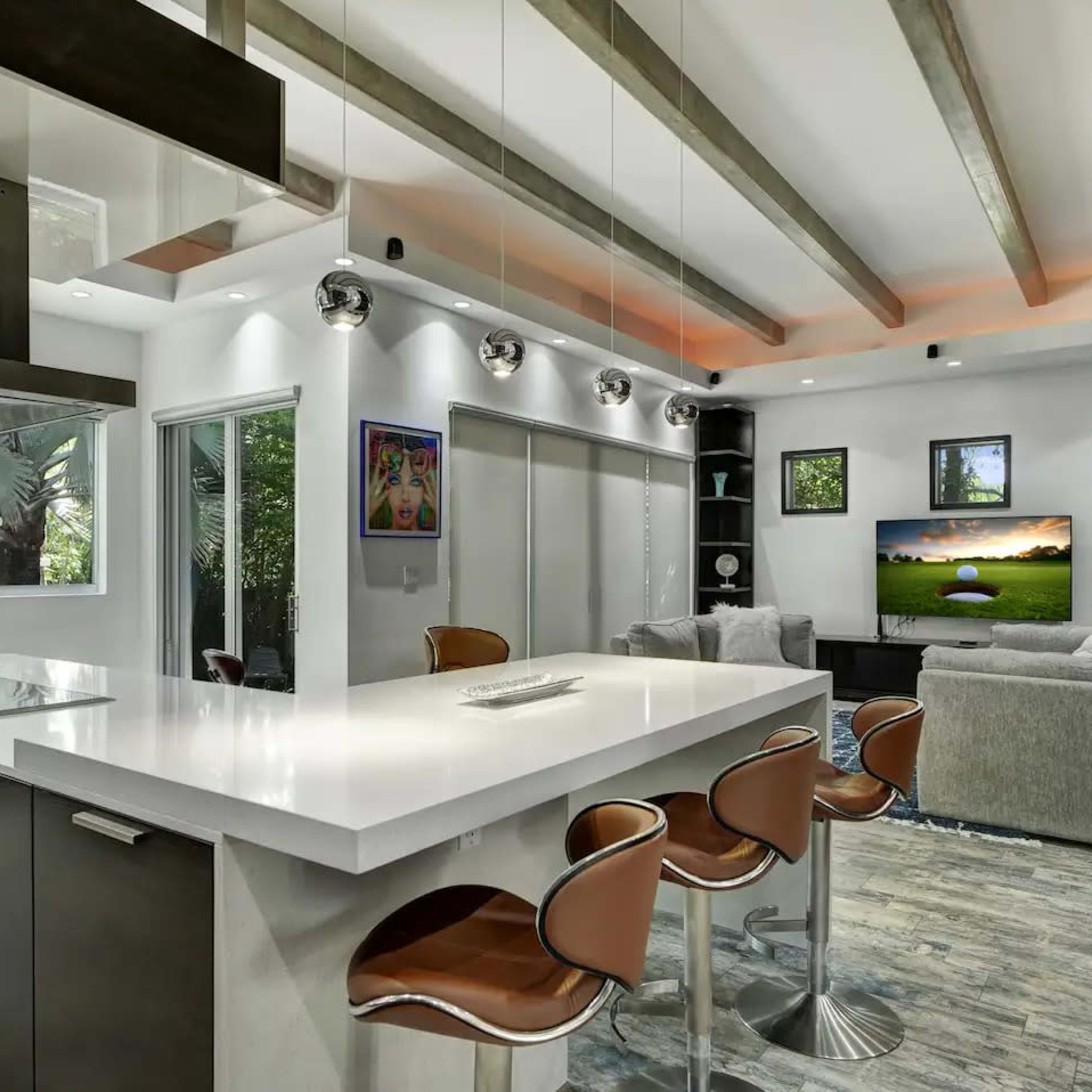 A modern kitchen and living area feature a large island with seating, sleek cabinetry, and large windows overlooking greenery.
