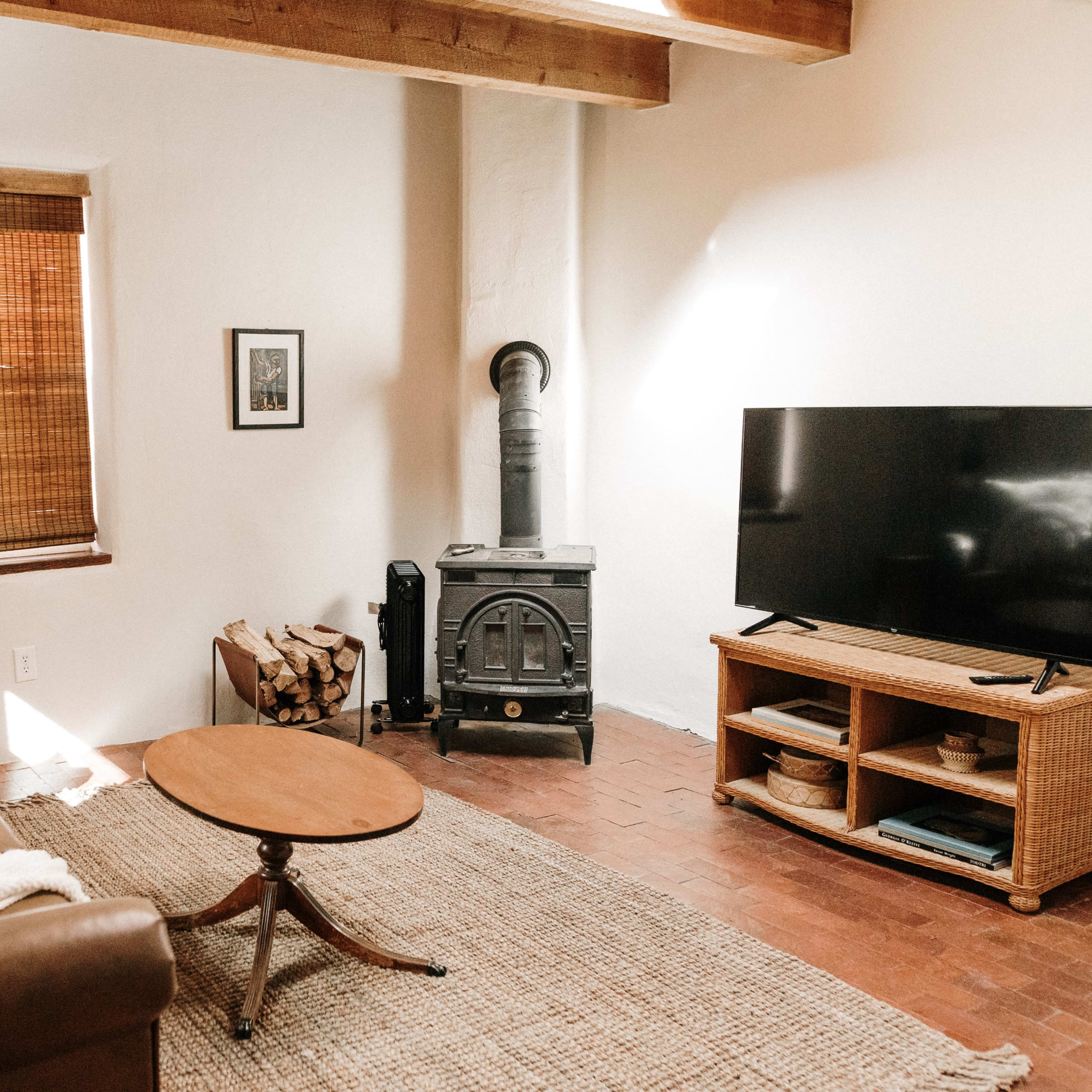 A cozy living room features a brown leather couch, a wooden coffee table, a television on a stand, and a wood-burning stove beside stacked firewood.
