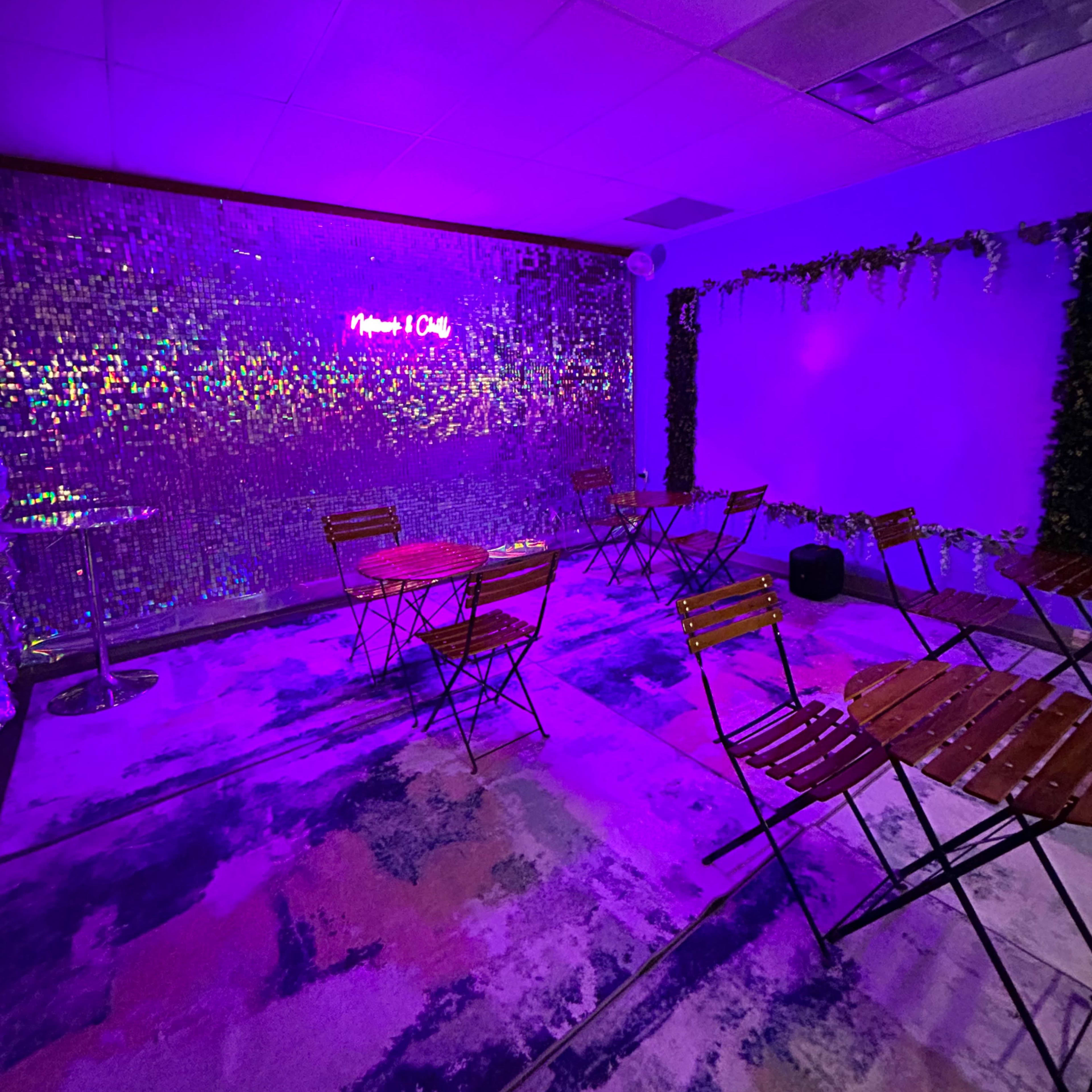 A small event space with purple lighting, featuring several wooden tables and chairs arranged on a colorful rug, with a shiny backdrop and a neon sign on the wall.