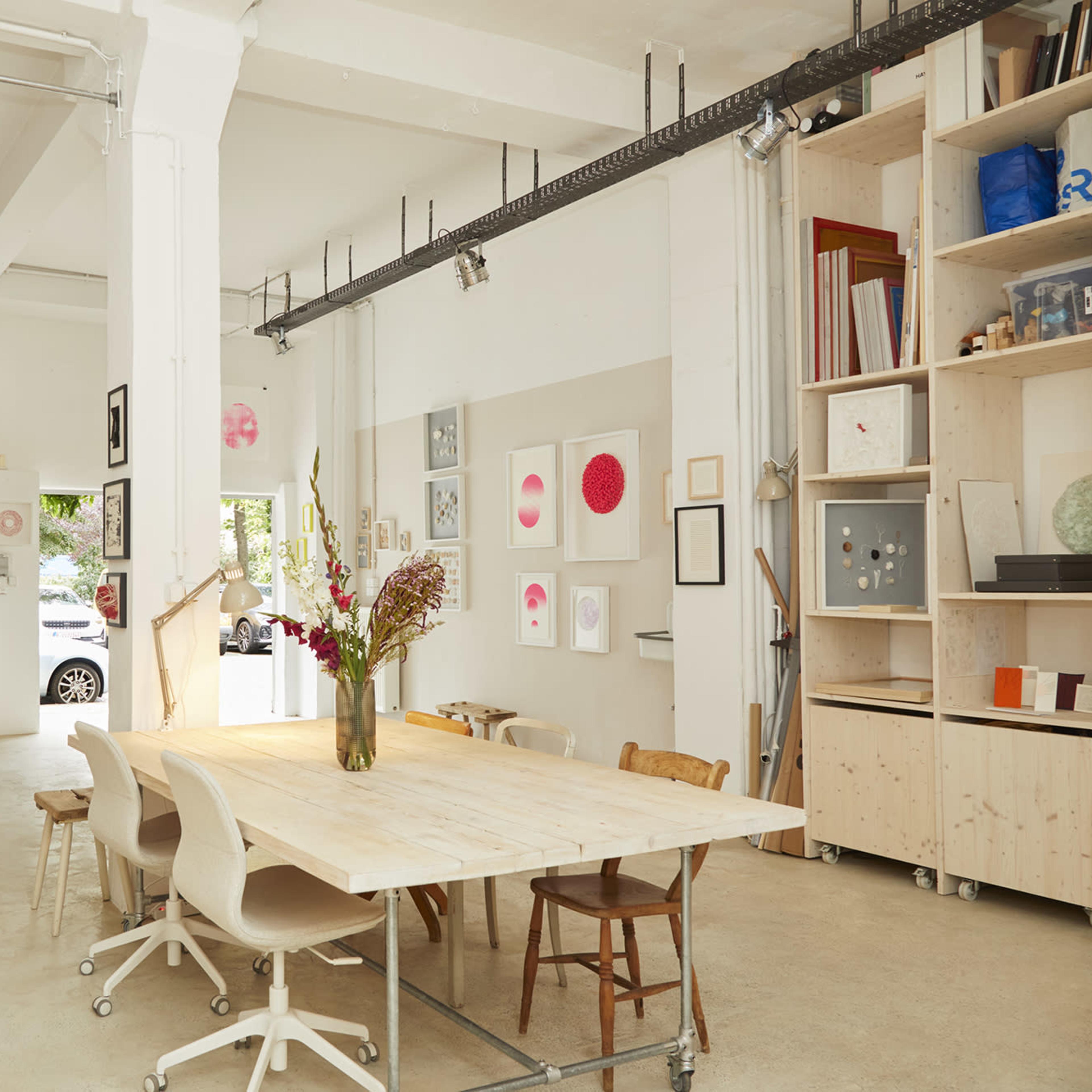 A bright, open studio space with wooden tables, white walls adorned with artwork, and shelves filled with various materials.