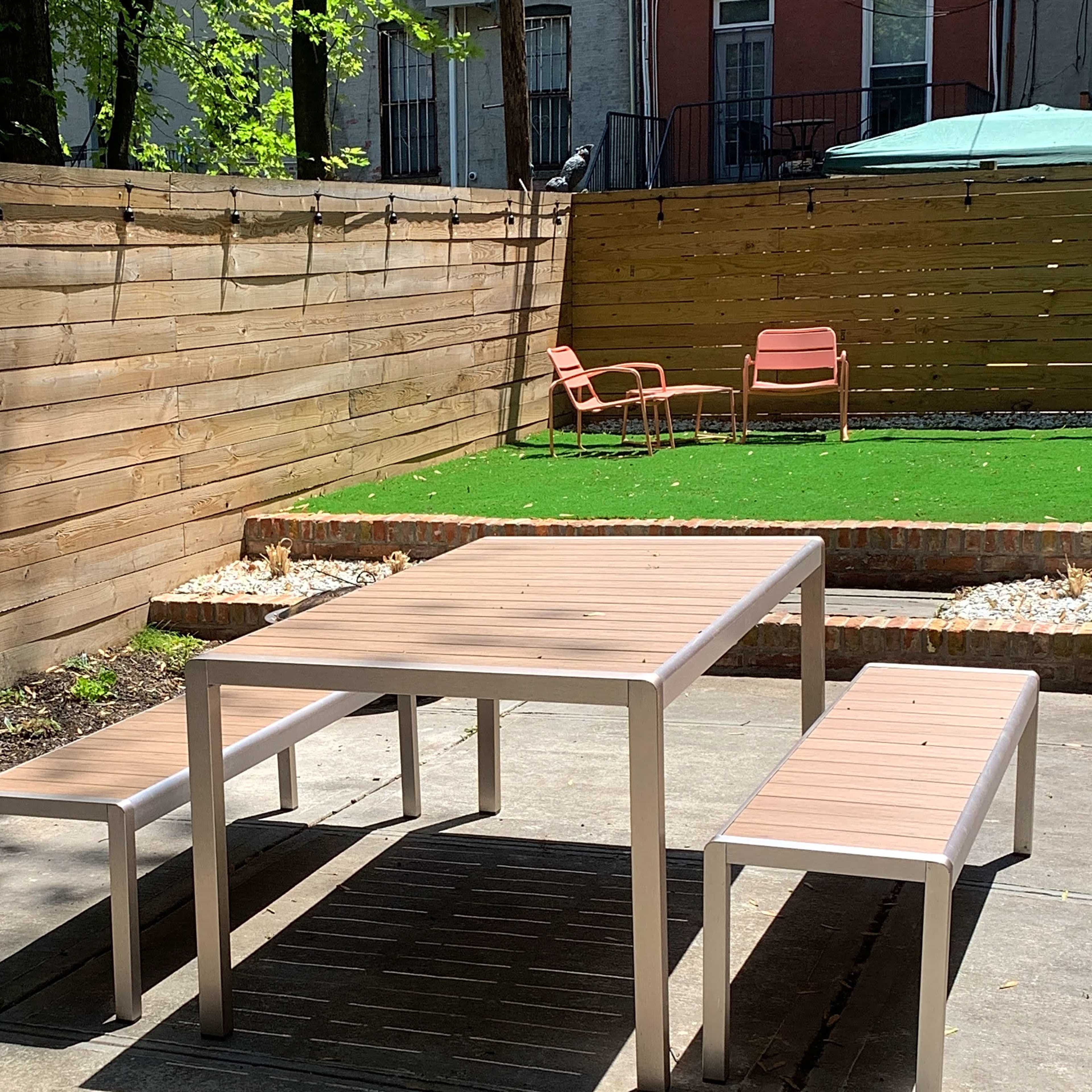 A patio area features a wooden fence, a table with benches, and two chairs on artificial grass.