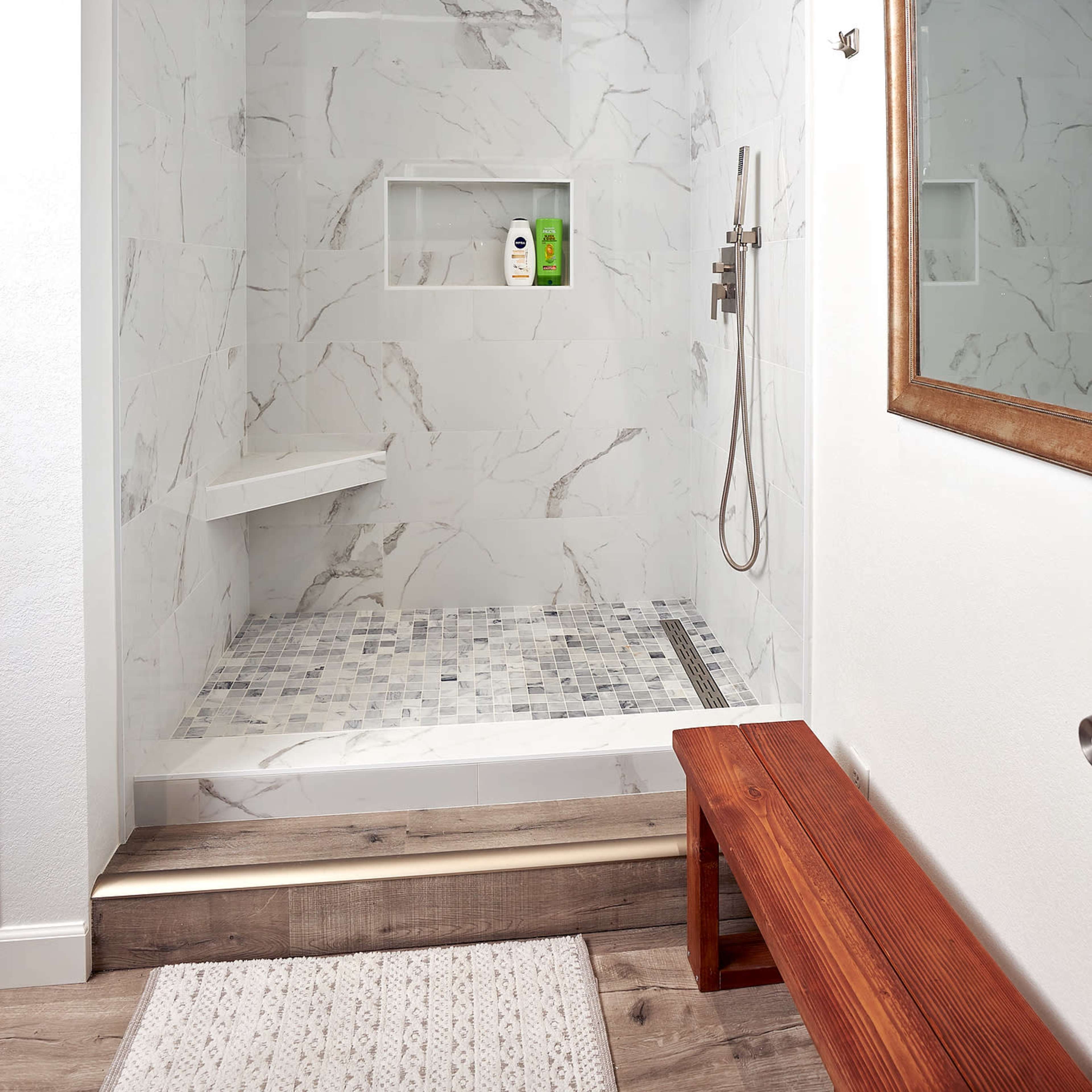 A modern shower stall with white marble walls, a glass partition, a built-in shelf for toiletries, and a wooden bench outside.