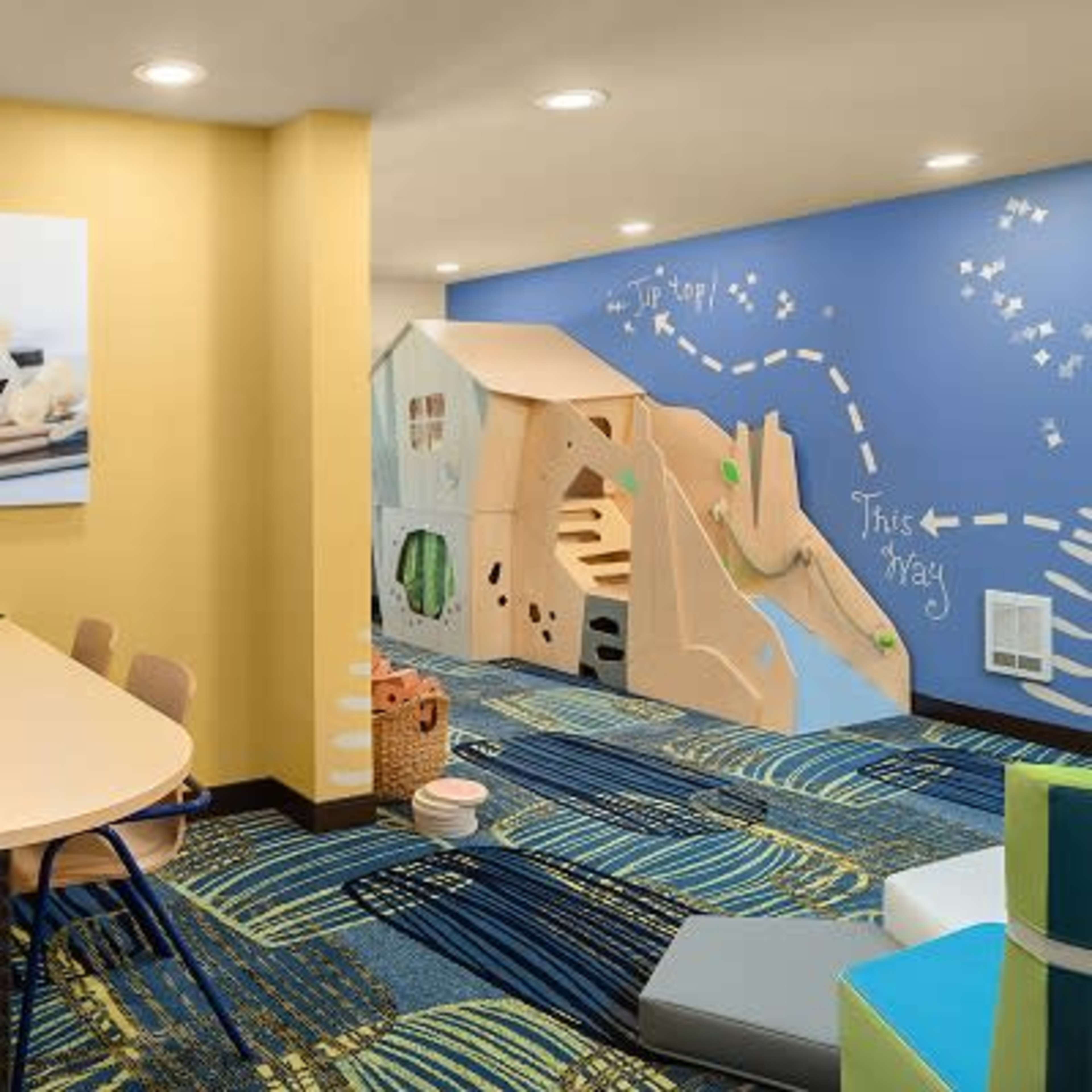 A colorful children's play area featuring a table with wooden toys and a playhouse structure against a wall with a blue mural and directional arrows.