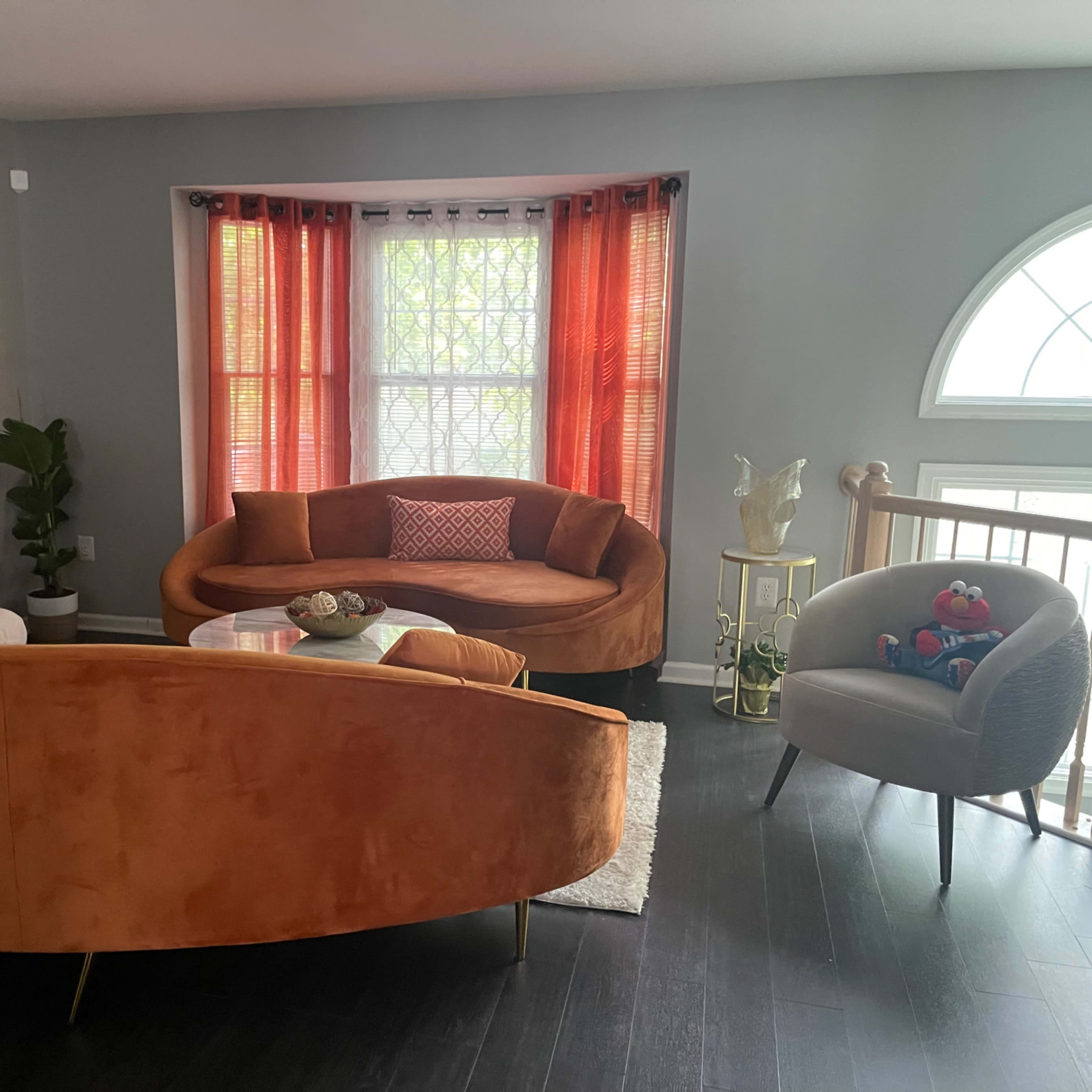 A modern living room with a circular orange sofa, a gray armchair, a small glass coffee table, and large windows draped with orange curtains.