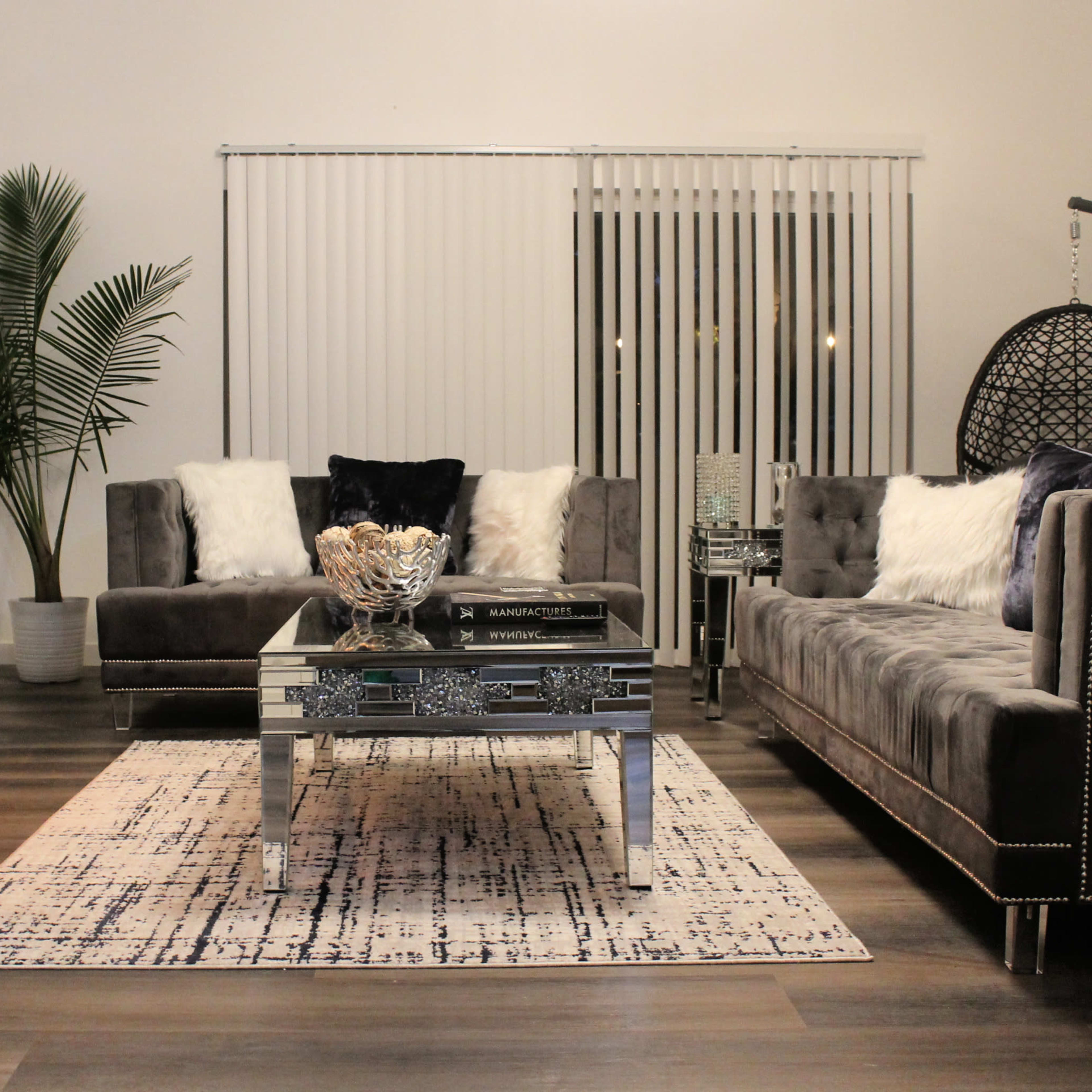 A modern living room features a gray sectional sofa, a mirrored coffee table, and a cozy fireplace, with a plant in the corner and large windows adorned with vertical blinds.