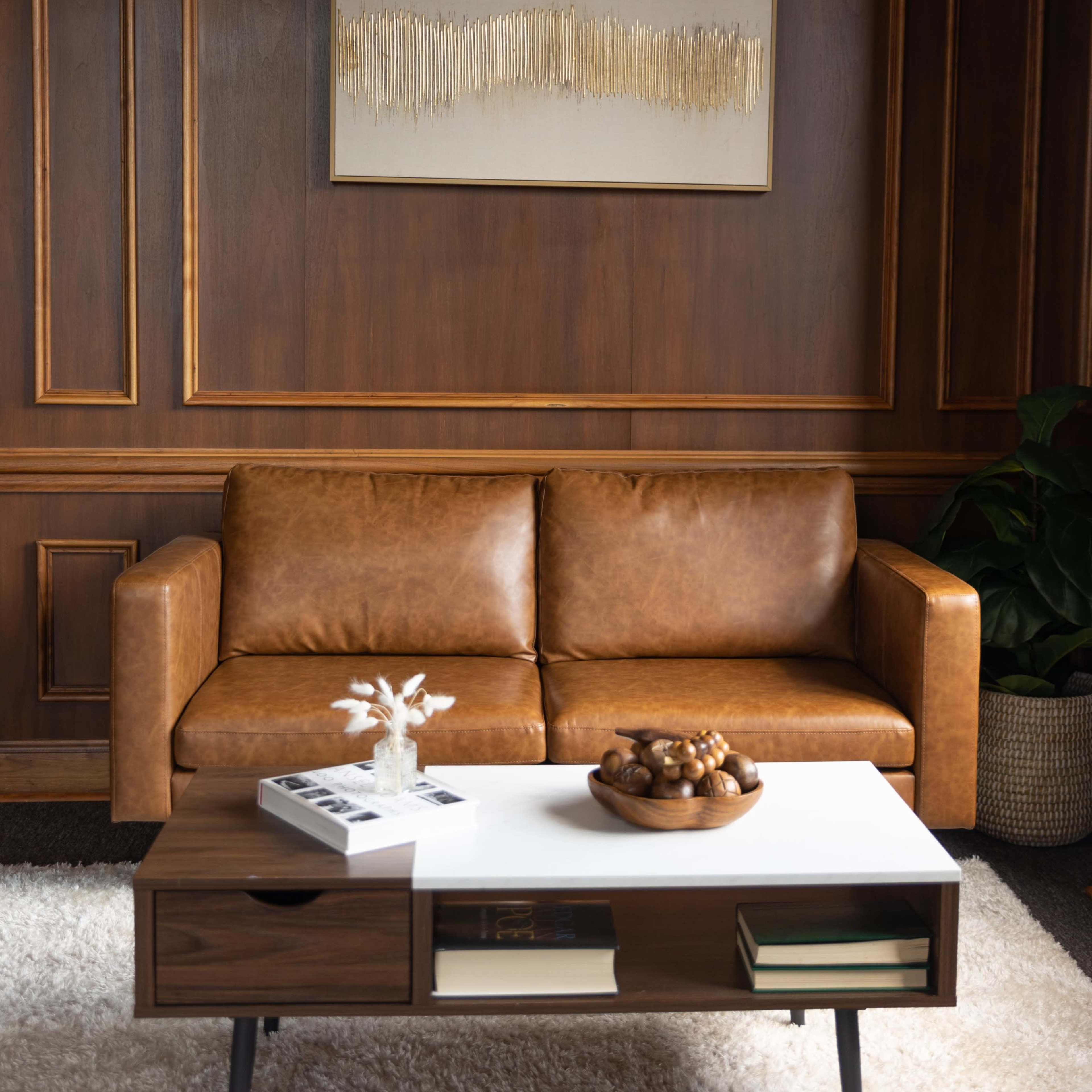 A brown leather couch sits in front of a wooden paneled wall, accompanied by a white coffee table adorned with decorative items and a textured artwork above it.