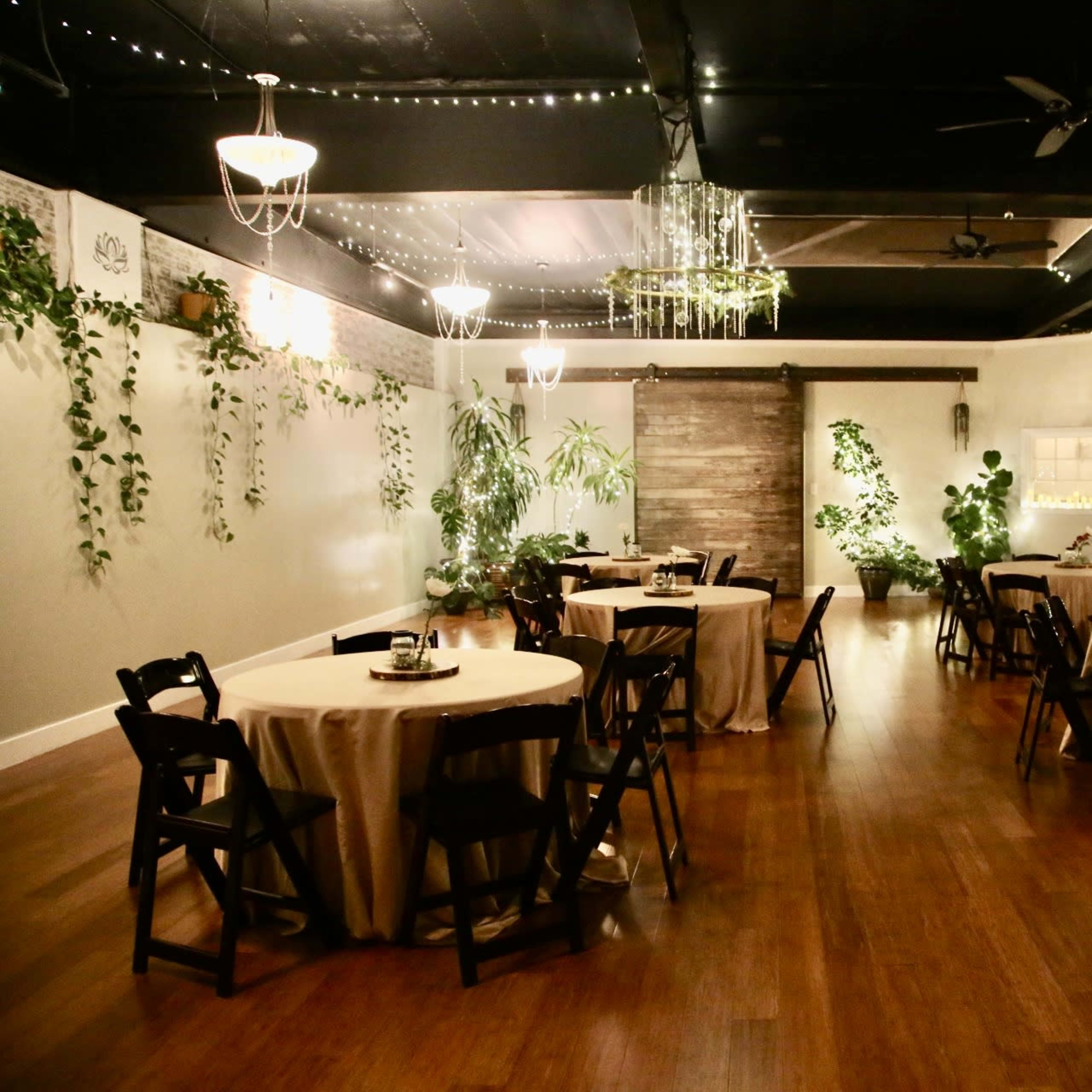 The room features several round tables covered with cream tablecloths, surrounded by black chairs, and is decorated with plants and dim lighting.