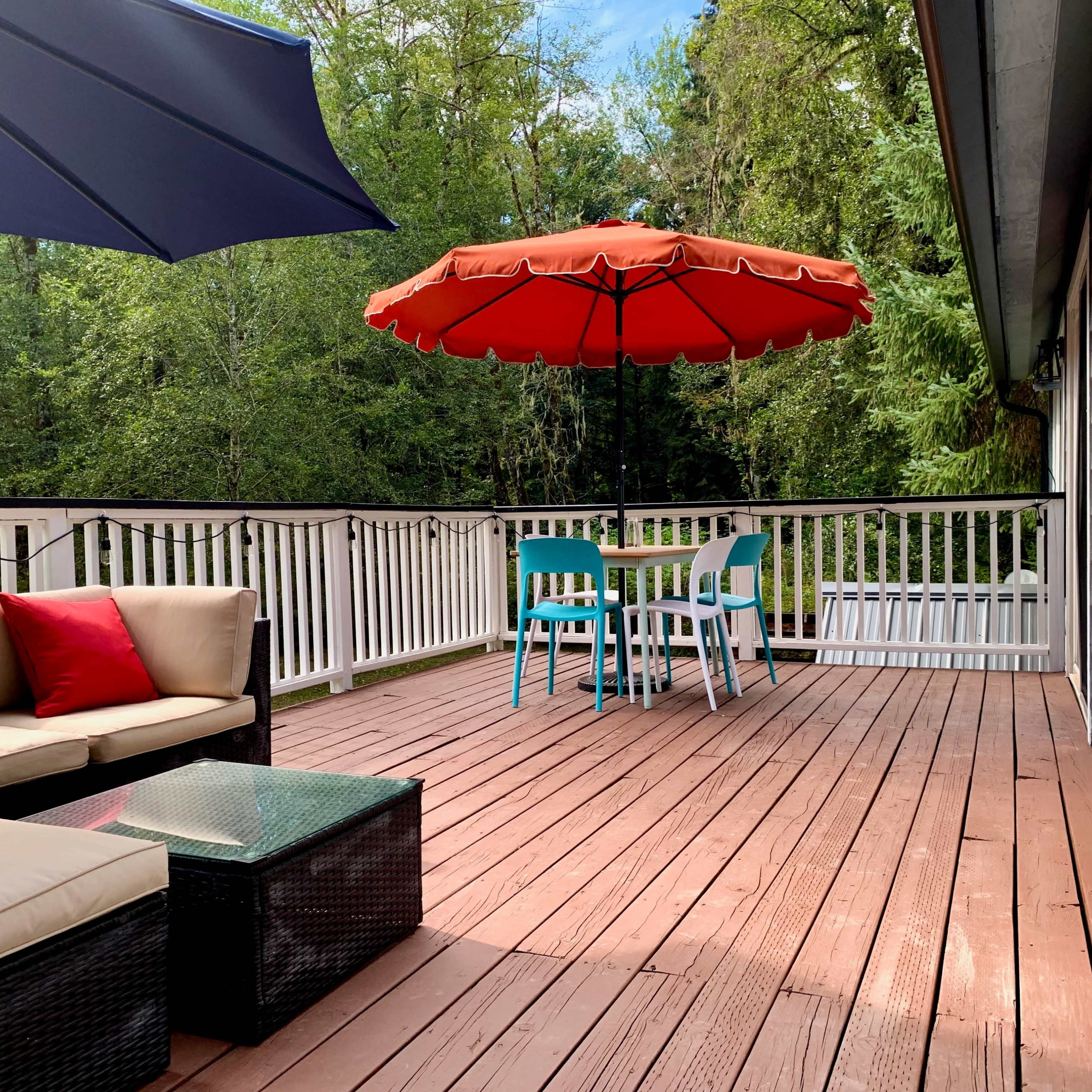 A wooden deck features a sofa with a red cushion, a glass coffee table, and a dining set shaded by an orange umbrella, surrounded by greenery.