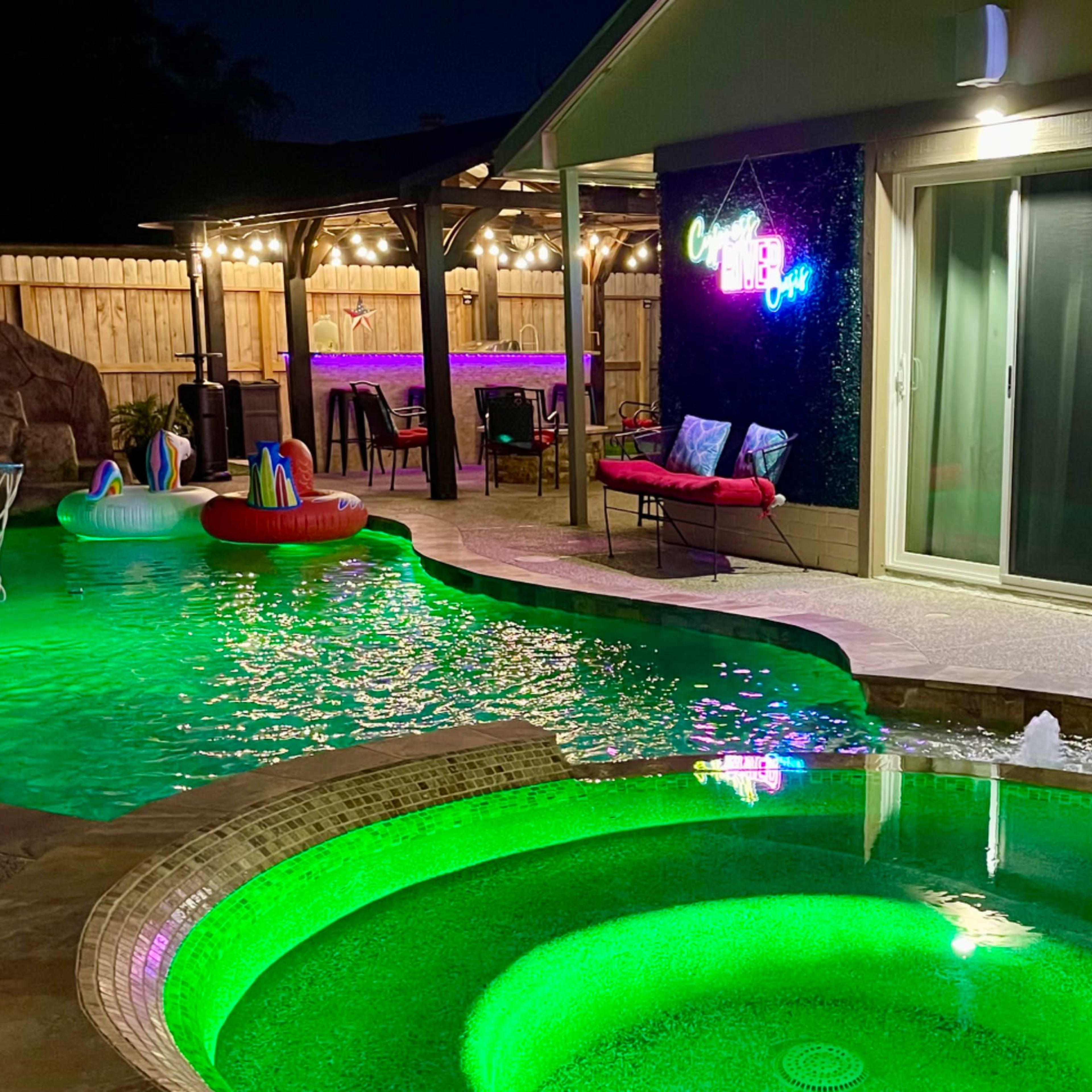 A backyard scene featuring a glowing pool with a spa, a TV mounted on a wall displaying a sports game, and seating areas illuminated by string lights.