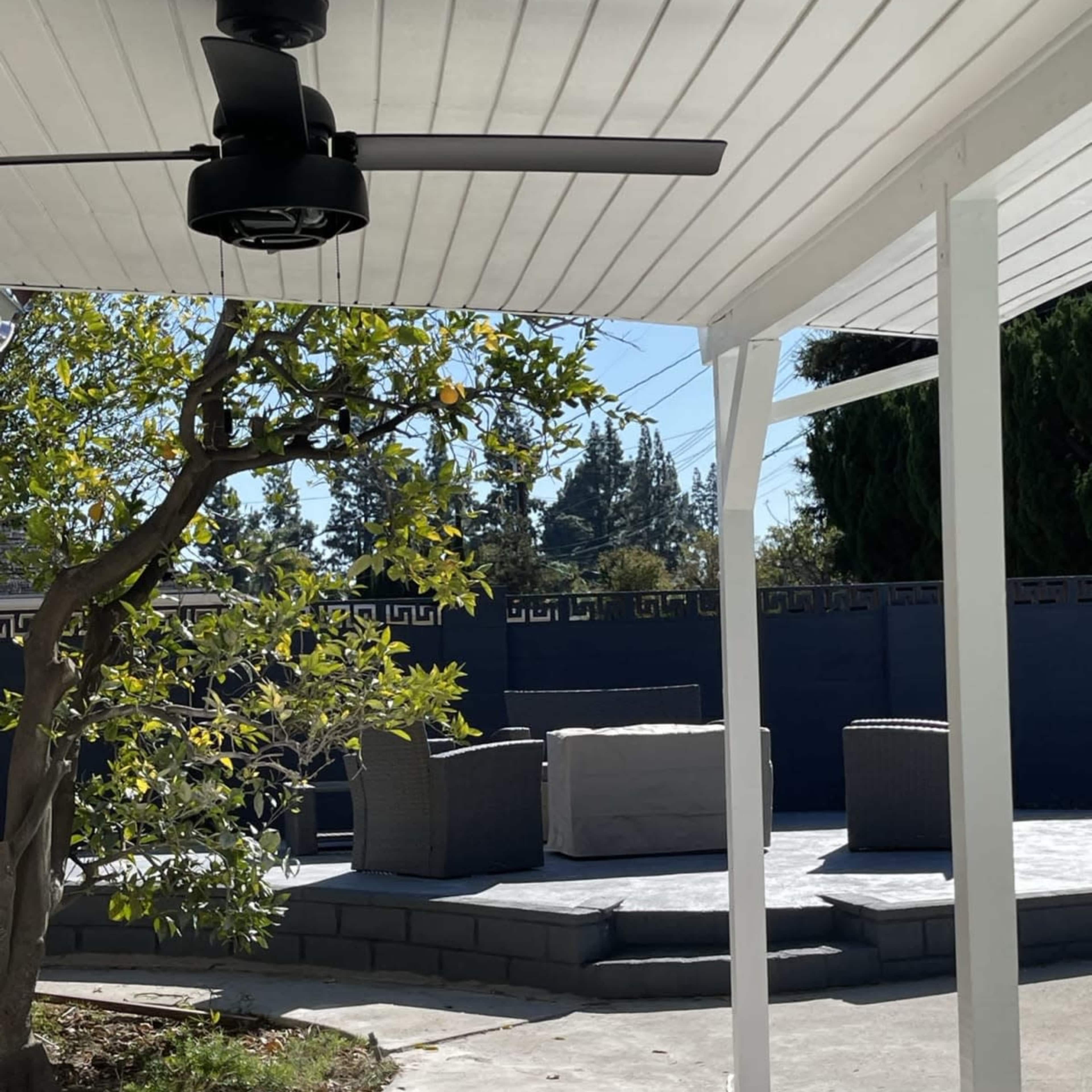 A patio area with a ceiling fan, featuring outdoor furniture arranged on a stone platform surrounded by greenery.