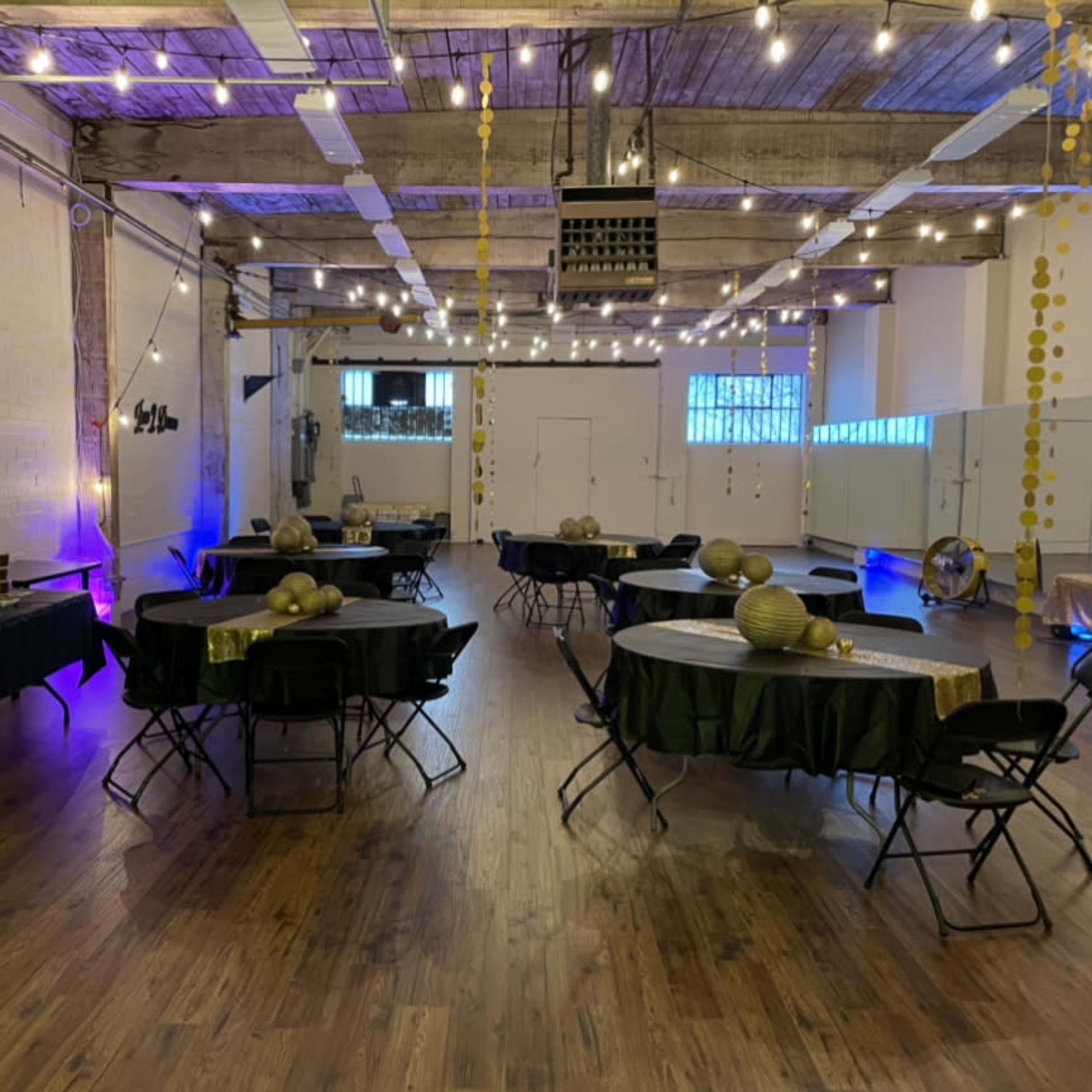 A spacious event hall set up with round tables covered in black and green tablecloths, illuminated by string lights and downlighting.