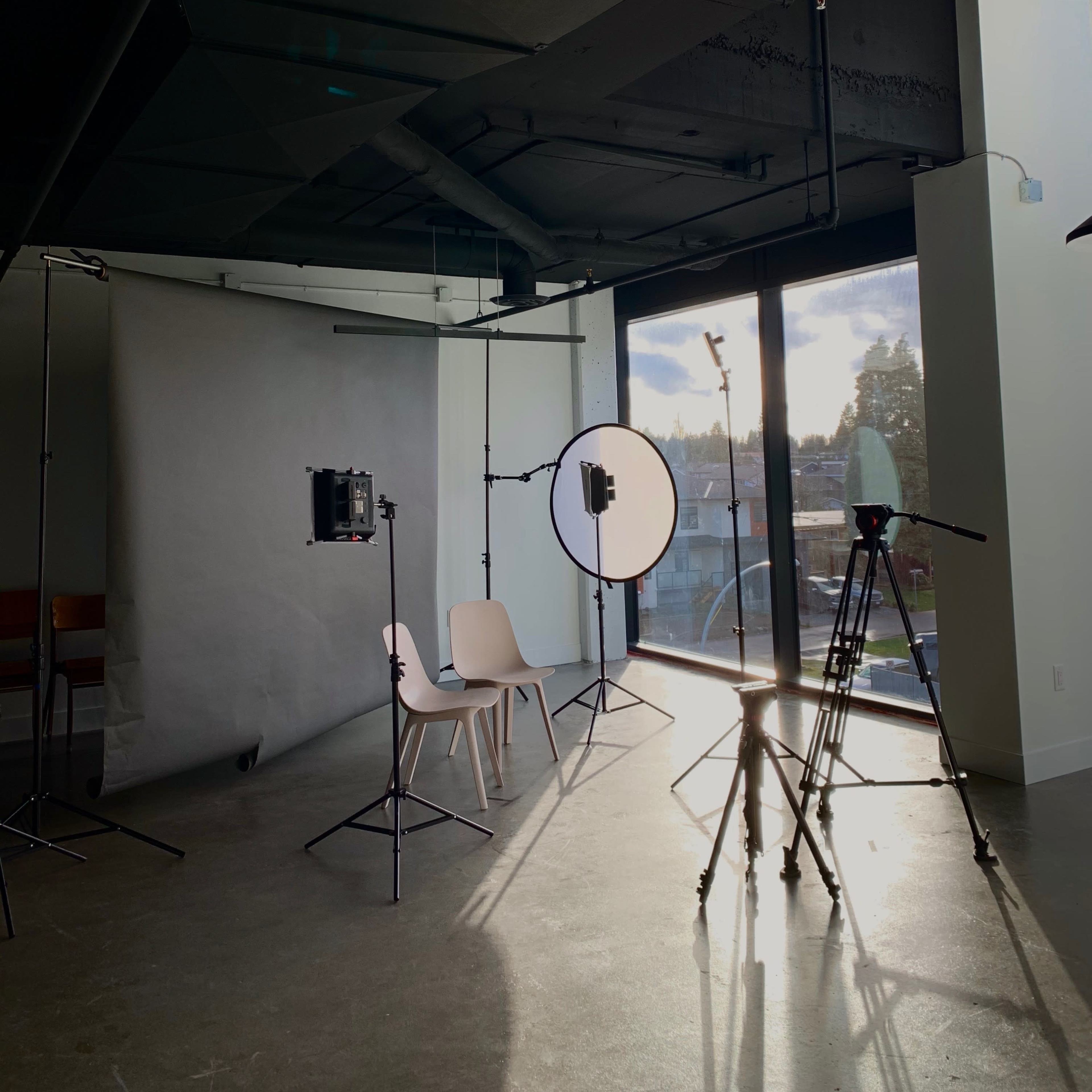 A photography studio with equipment, including a chair, light stands, a circular reflector, and a large window letting in natural light.