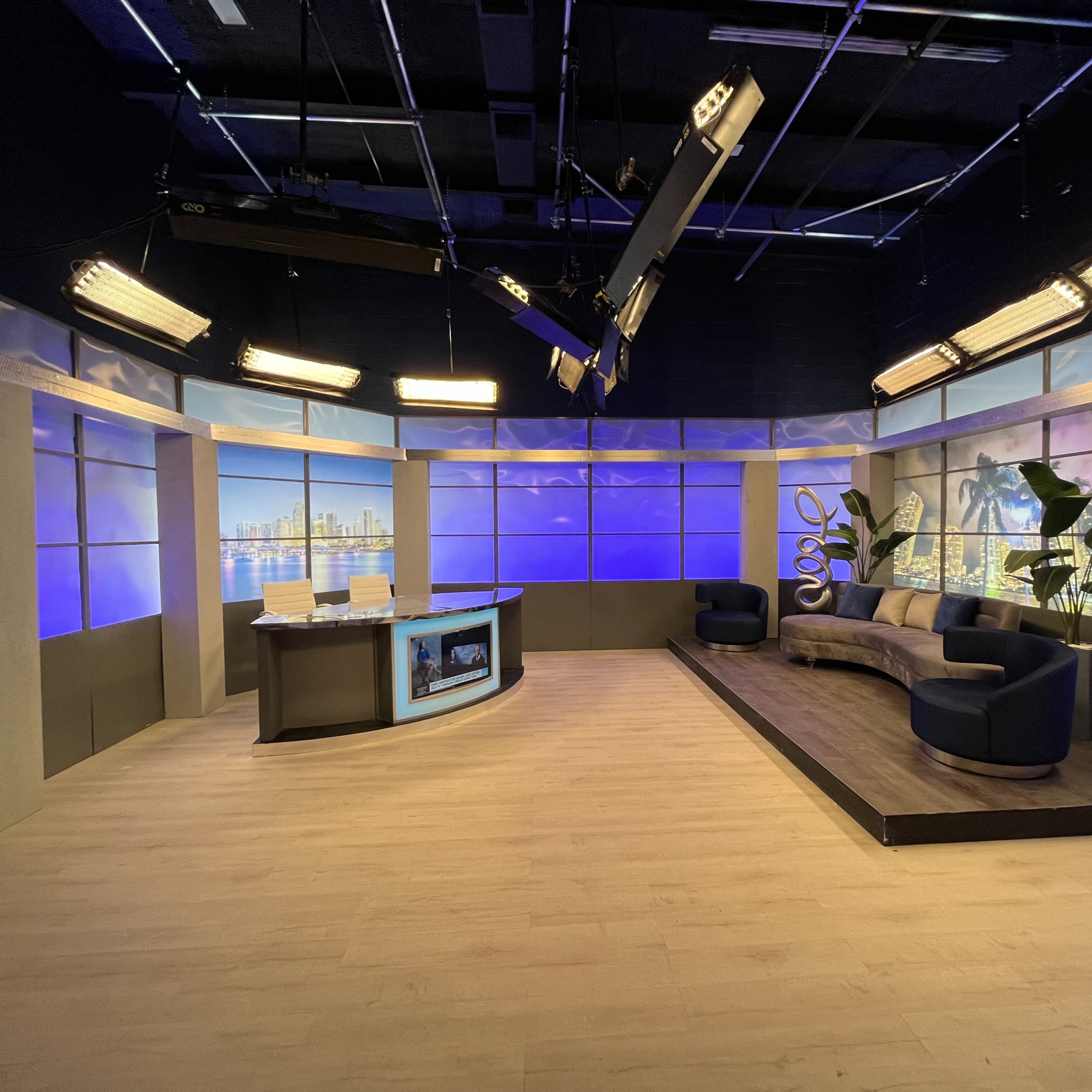 A modern television studio features a newsroom desk, a seating area with a sofa, and large windows displaying a city skyline backdrop.