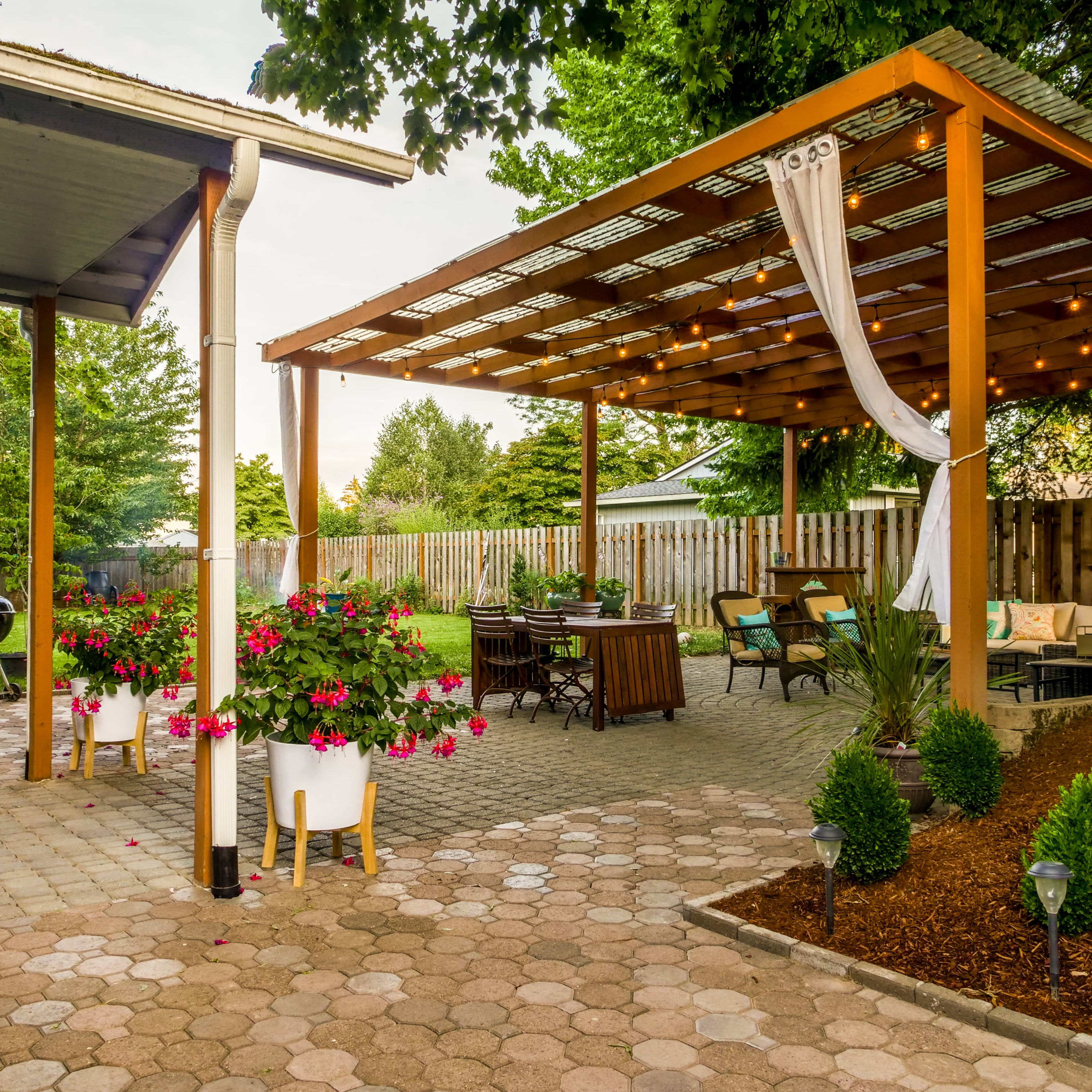 A backyard patio with a wooden pergola, outdoor seating, hanging lights, and flower pots next to a paved walkway.