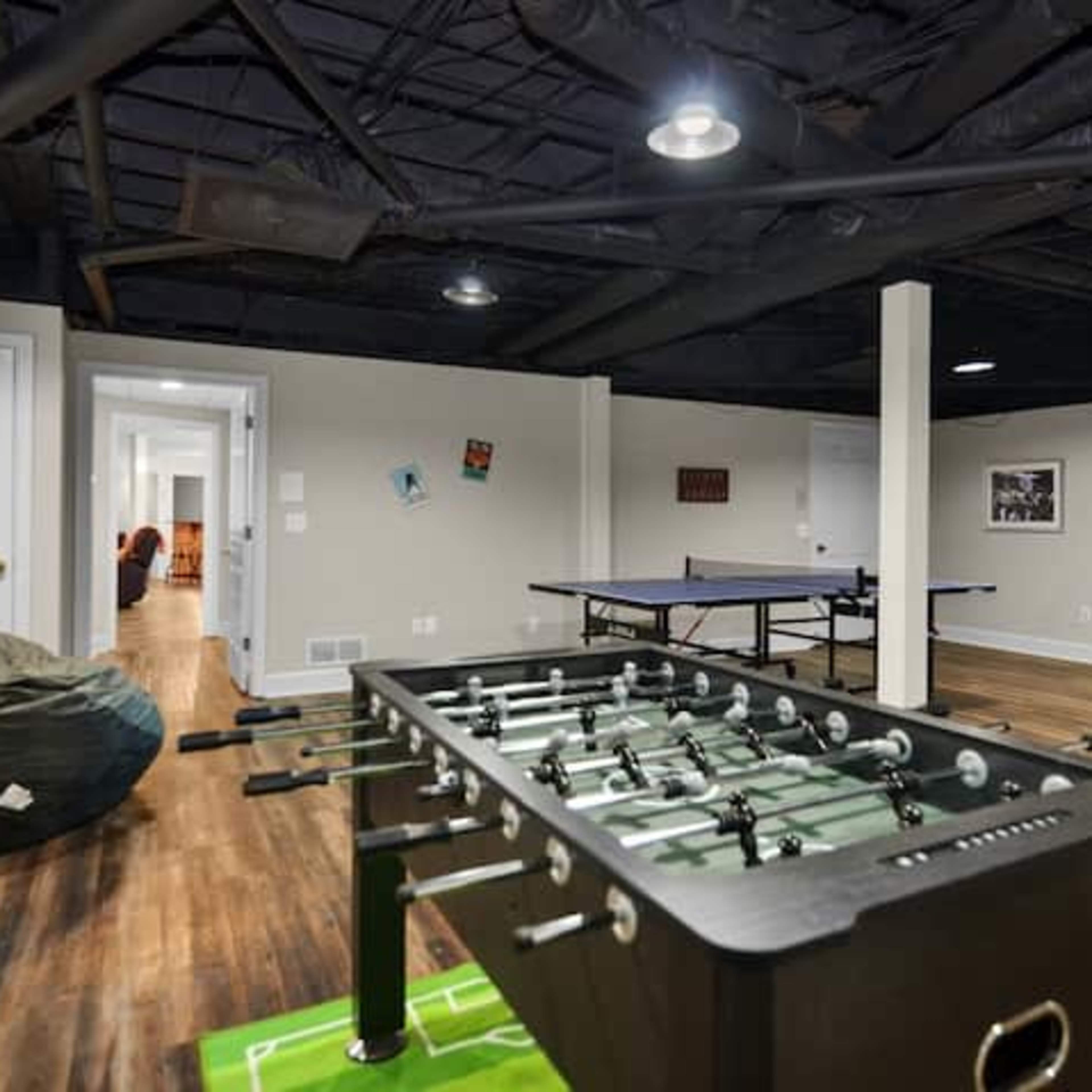A spacious game room featuring a foosball table, ping pong table, and bean bag chairs, with a hallway leading to another room in the background.